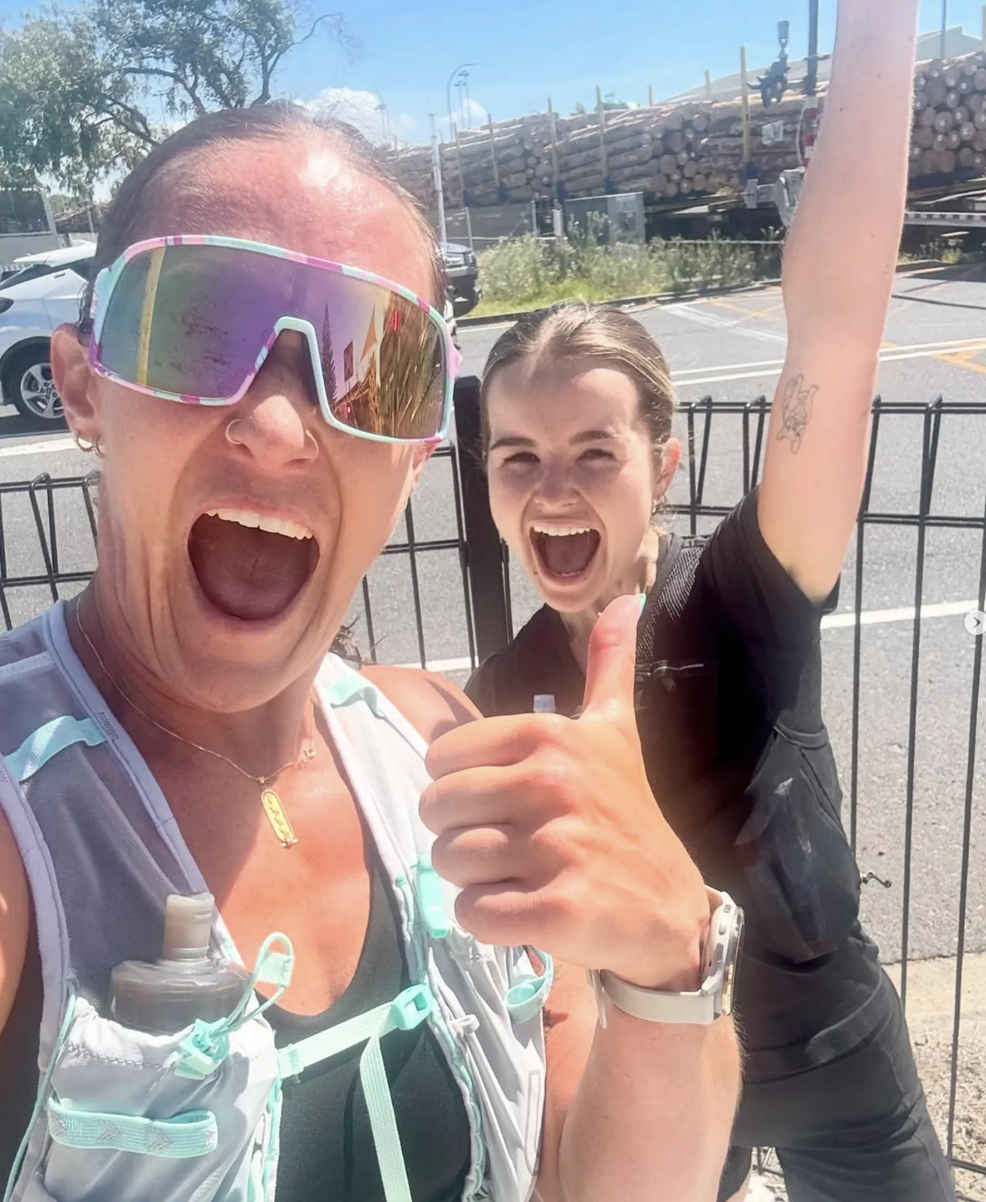 Two women in athletic gear smile and cheer outdoors on a sunny day. One gives a thumbs up and wears reflective sunglasses; the other raises her arm excitedly. Logs and a fence are visible in the background.
