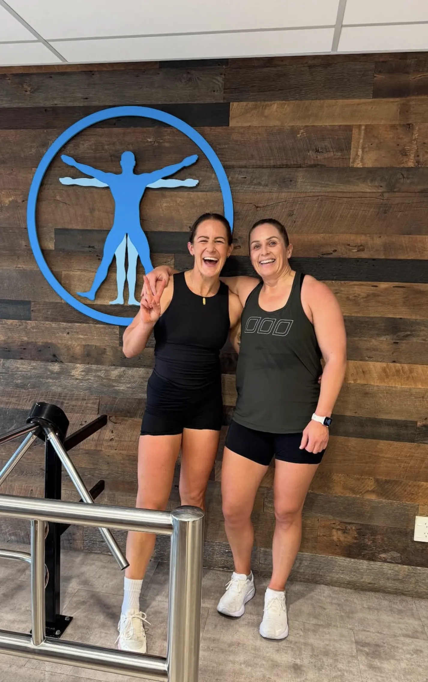 Two women in athletic wear smile and pose together in front of a wooden wall with a blue human figure logo. One woman holds up a peace sign, and both appear happy and energized in a gym setting.