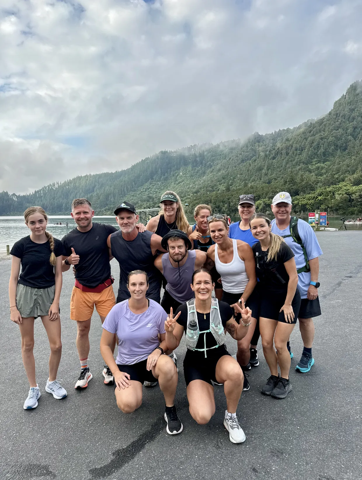 A group of thirteen people in athletic wear pose and smile for a photo in front of a misty lake and green forested hills under a cloudy sky. Some are standing, others kneeling, enjoying an outdoor activity.