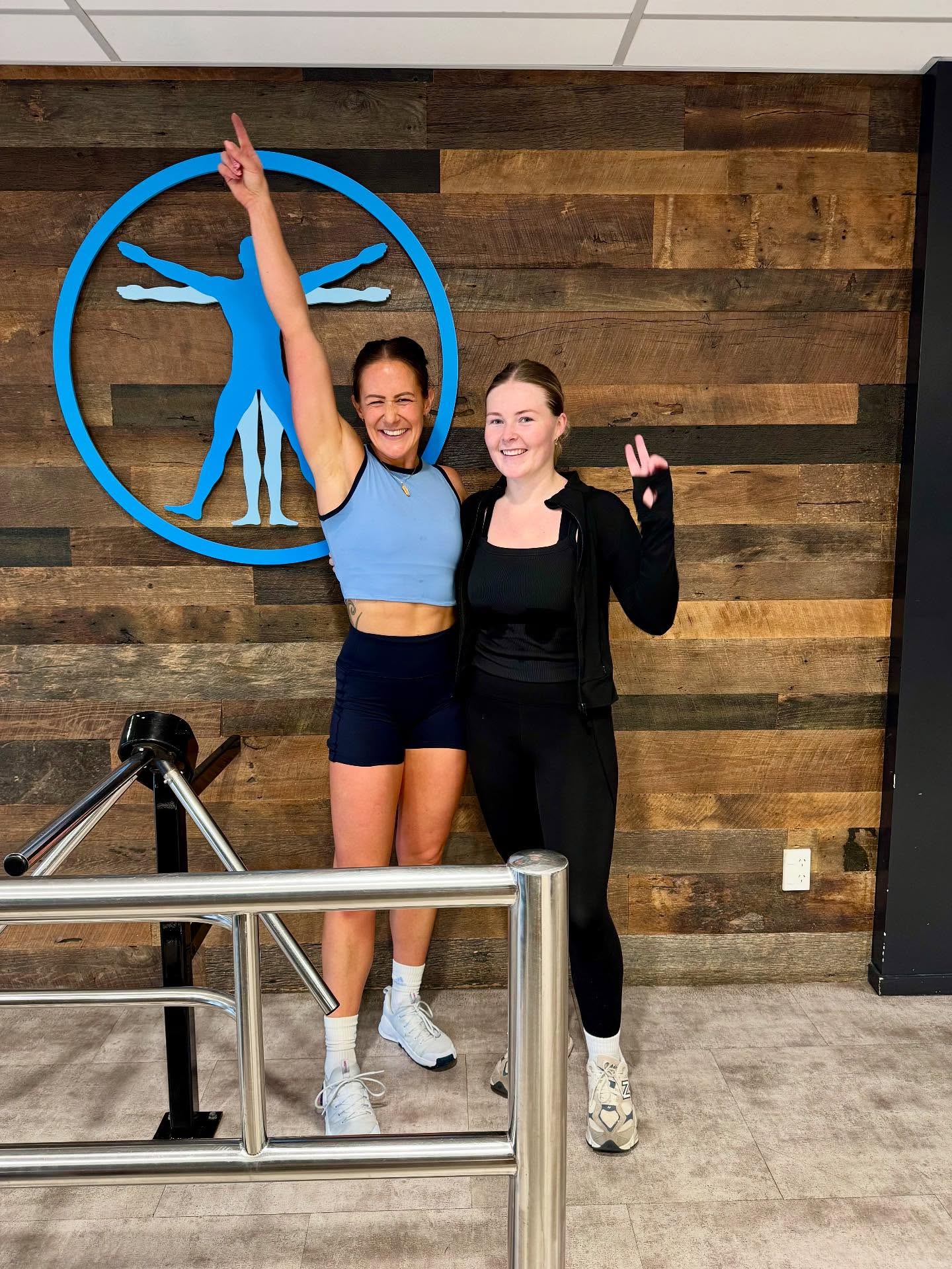 Two women pose and smile in front of a wooden wall with a blue human figure logo. One woman wears a blue crop top and shorts, raising an arm in celebration, while the other wears black athletic wear and flashes a peace sign.