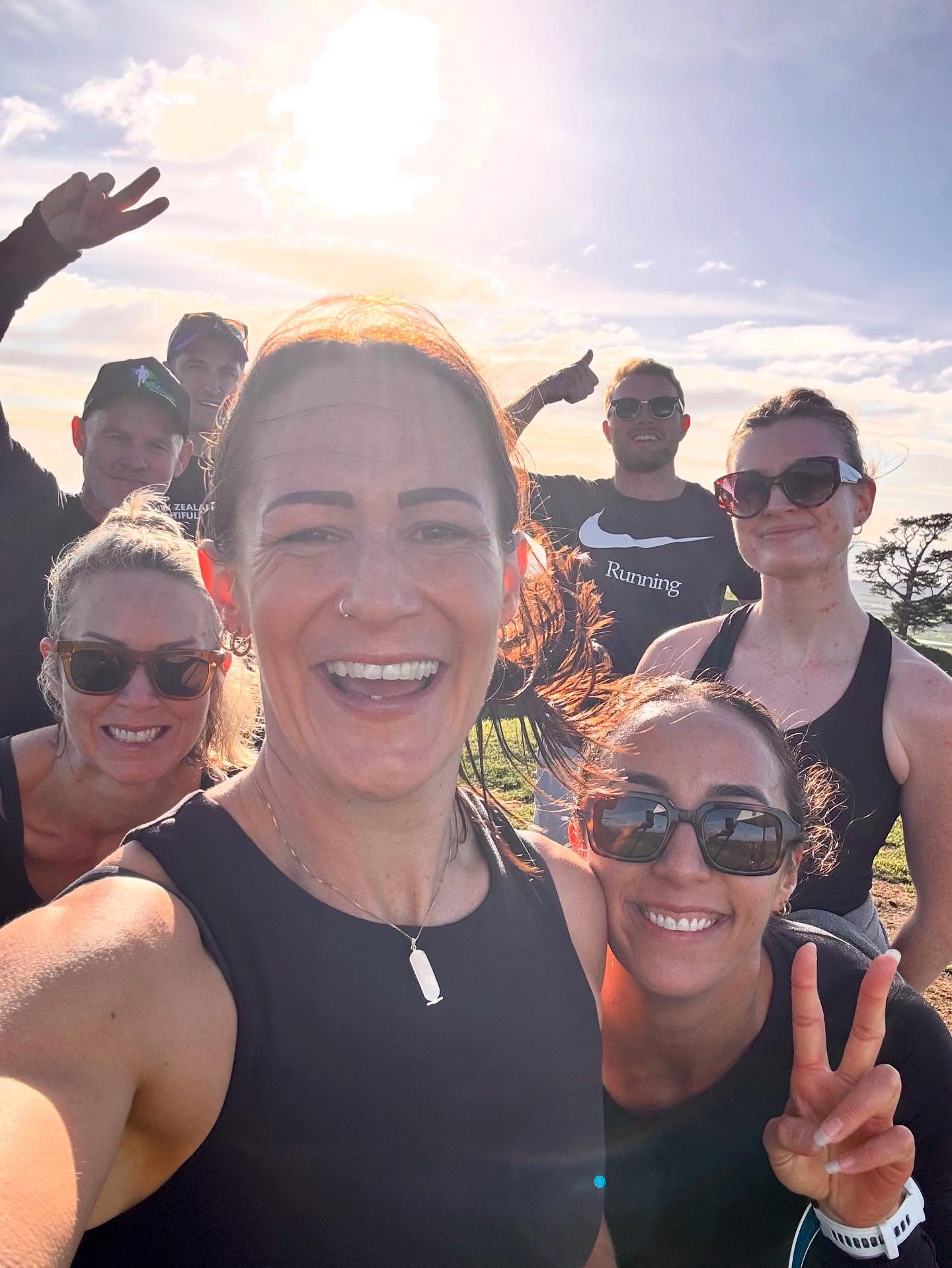 A group of smiling people in athletic gear take a selfie outdoors on a sunny day. Some are wearing sunglasses, and one person is making a peace sign. The sun is shining brightly in the background.