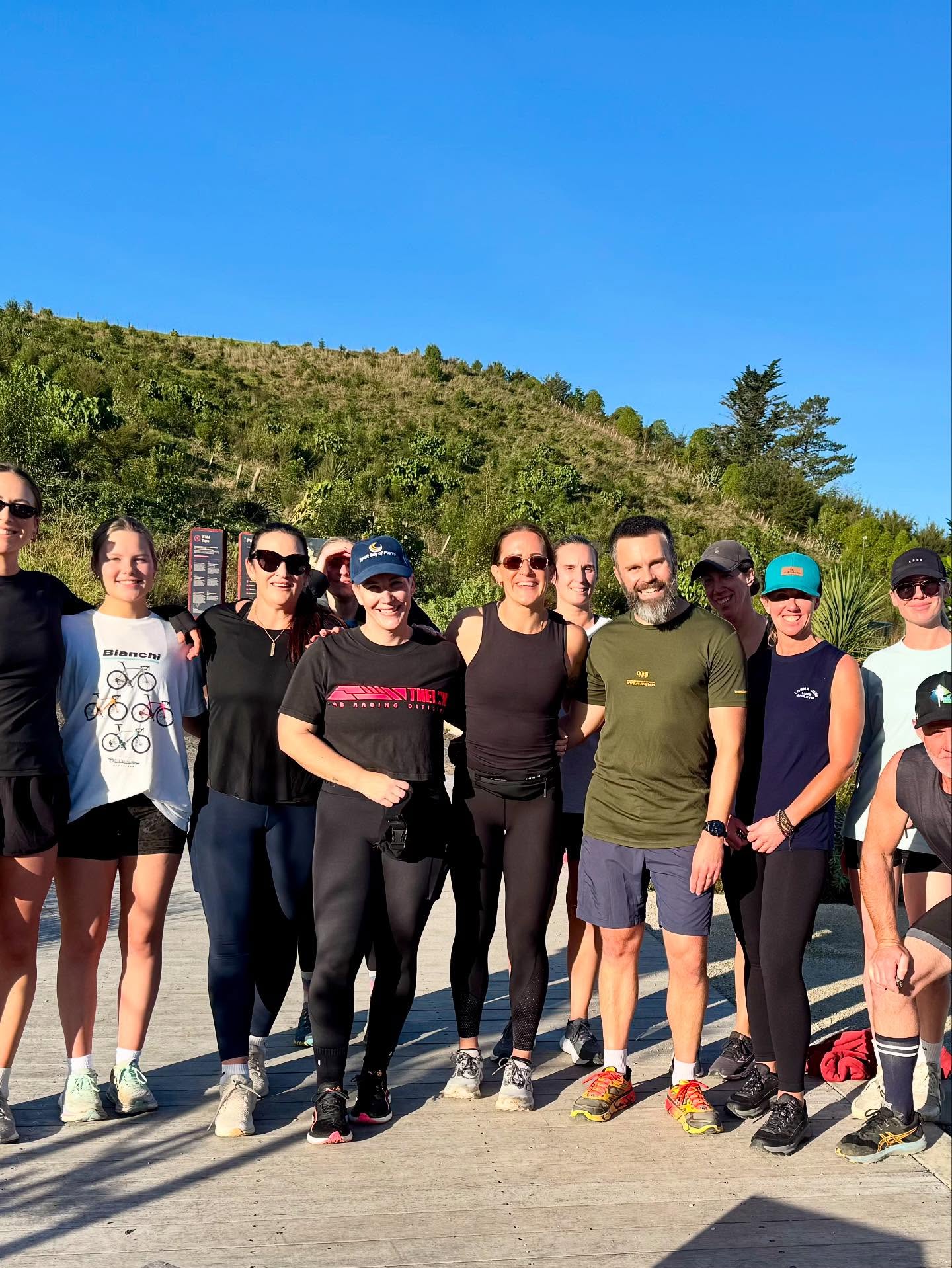 A group of people in athletic wear stand together outdoors, smiling at the camera, with trees and a grassy hill in the background under a clear blue sky.