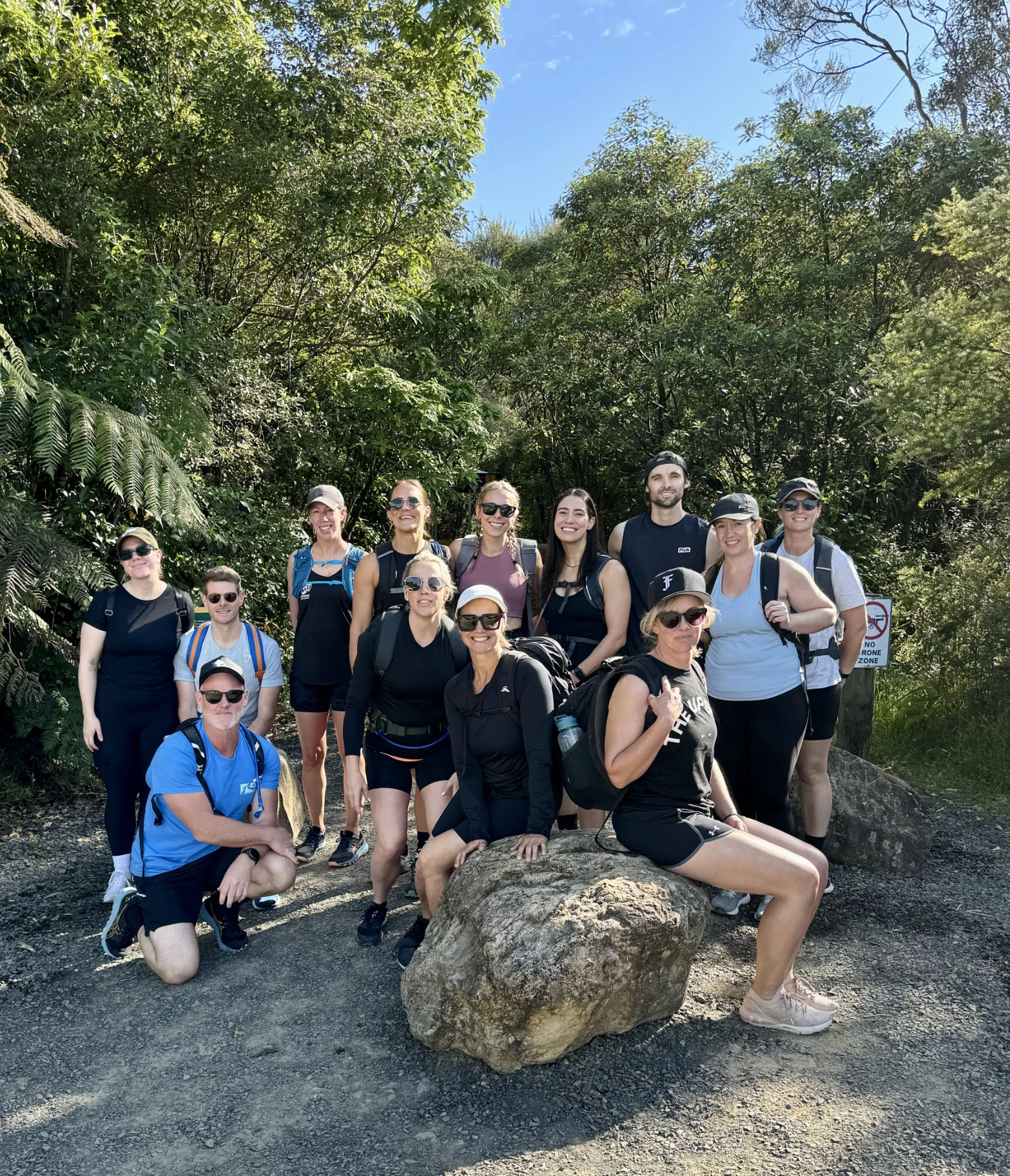 A group of 14 people pose outdoors on a trail, some standing and some sitting on a large rock, surrounded by green trees and bushes under a clear blue sky. Most are wearing sunglasses, hats, and casual hiking attire.