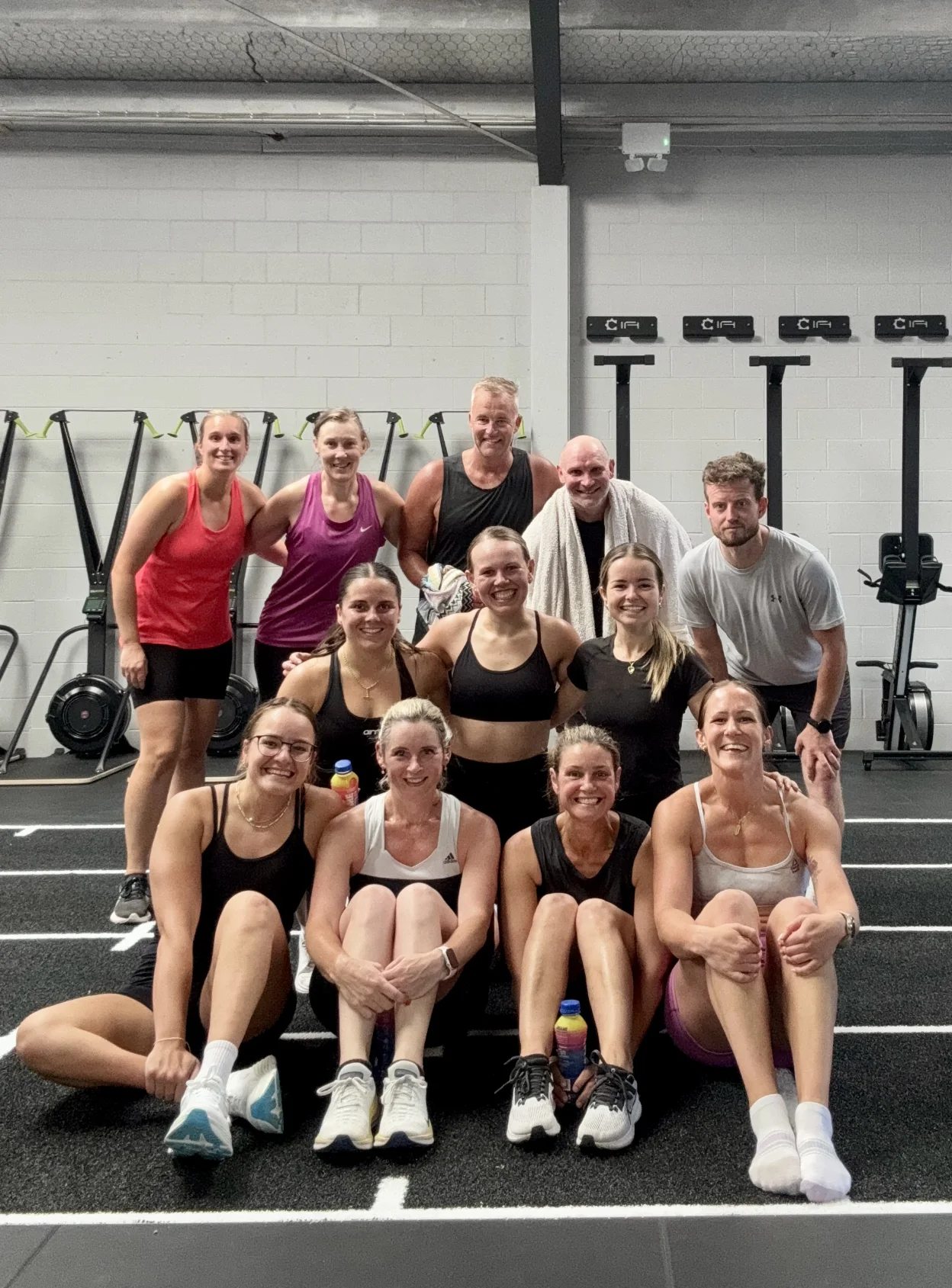 A group of thirteen adults in workout clothes sit and stand together, smiling at the camera in a gym with exercise equipment visible in the background.