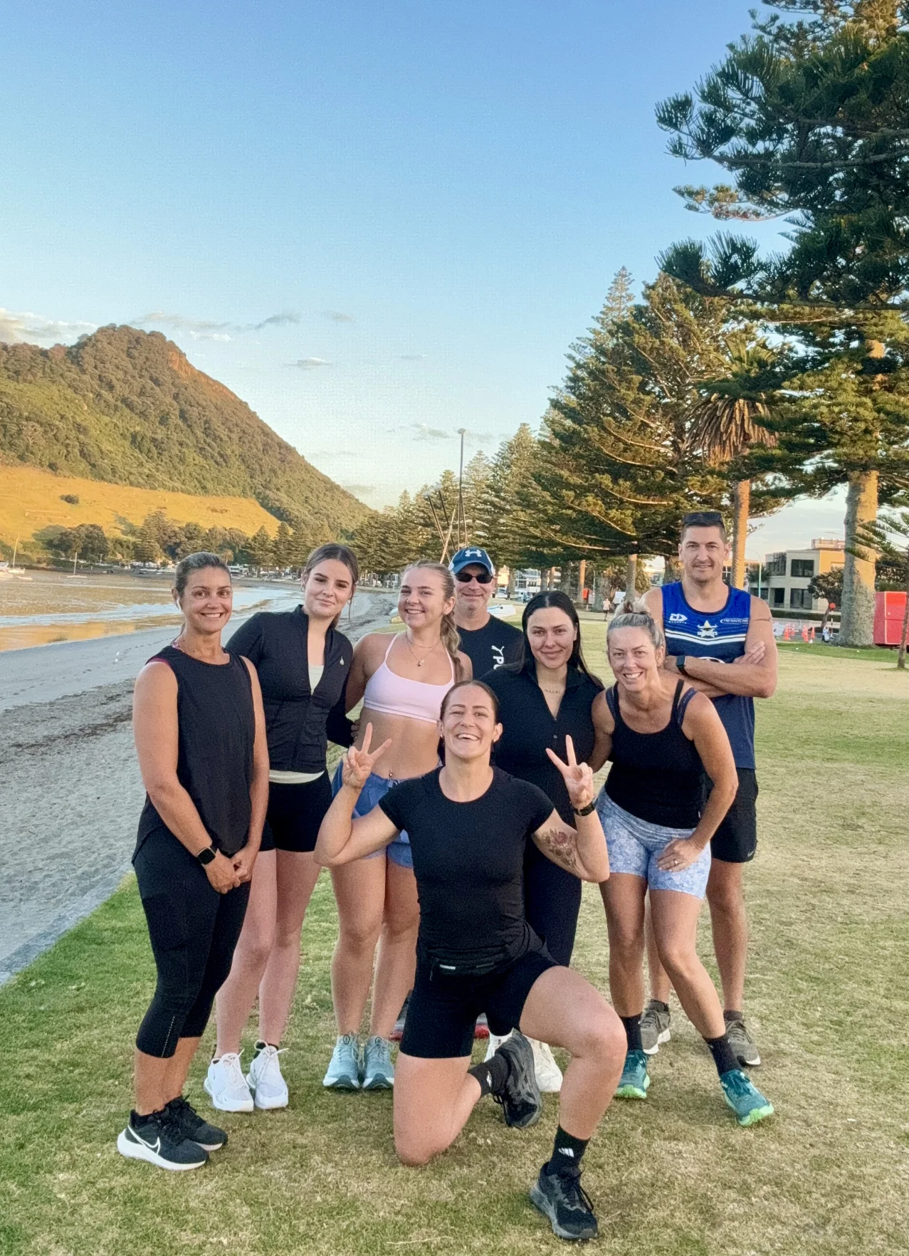 A group of eight people pose and smile outdoors on grass near a beach, with pine trees, a hill, and buildings in the background. One person in front is kneeling and making a peace sign. The weather is clear and sunny.
