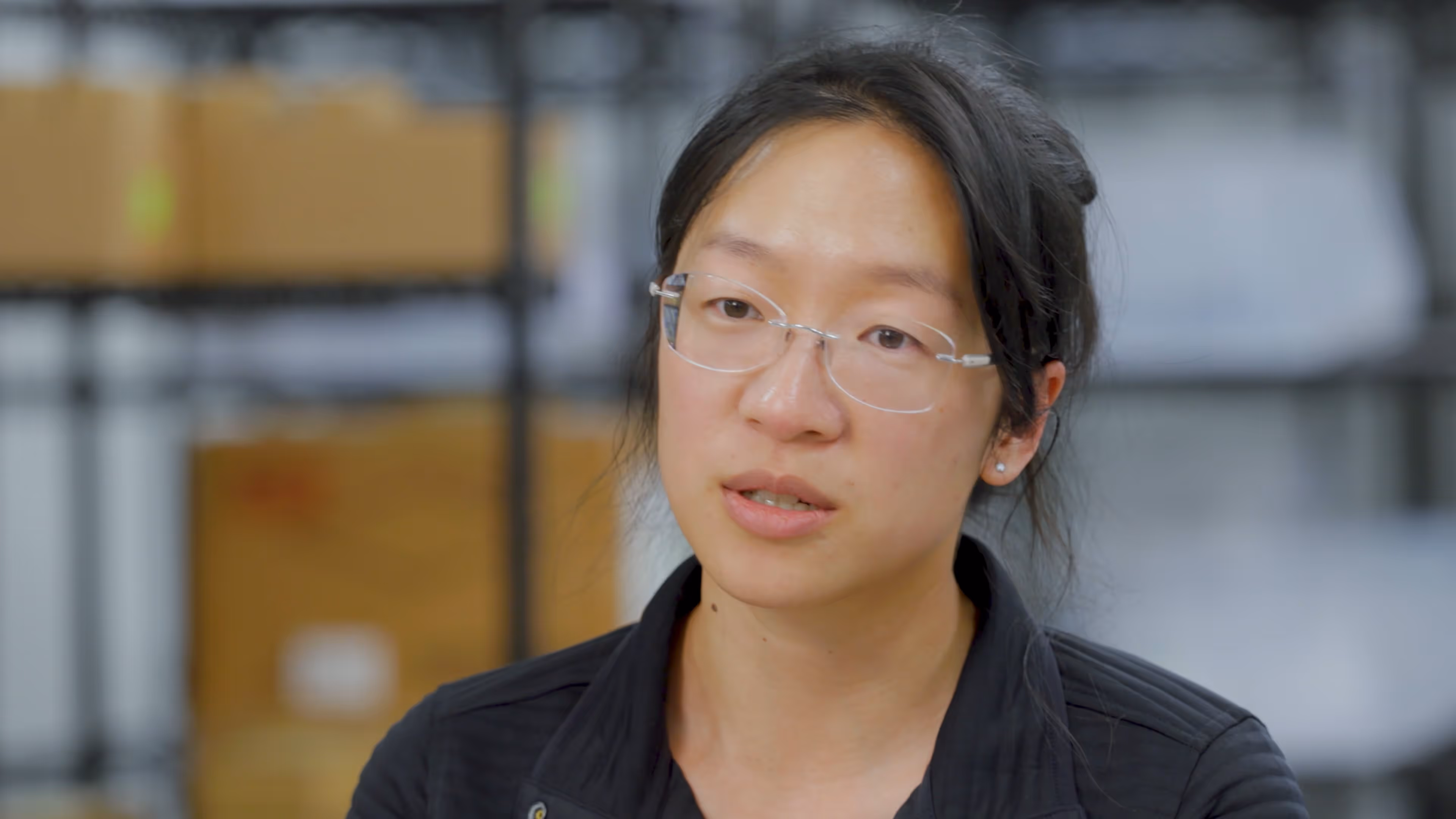Woman with glasses and dark hair pulled back wearing a black jacket in a warehouse setting.