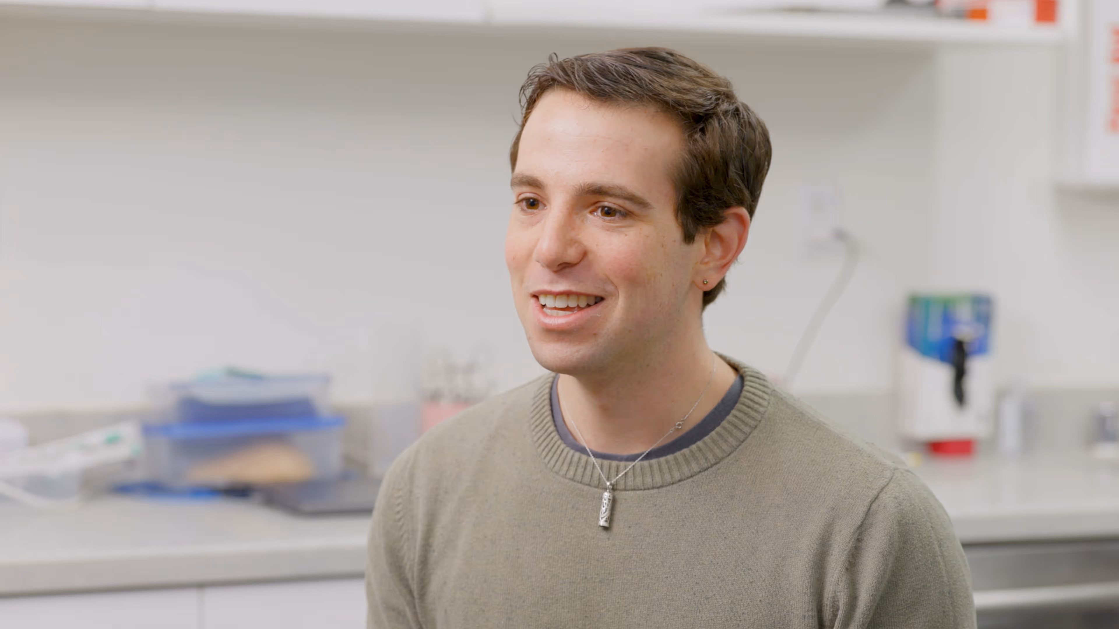Young man with short brown hair wearing a gray sweater and necklace smiling indoors.