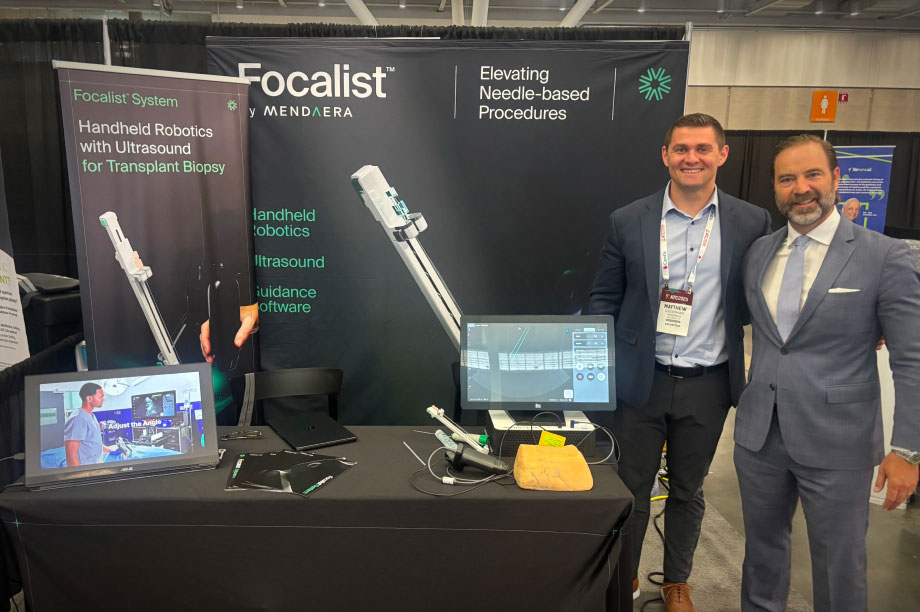 Two men in business attire stand smiling at a booth for Focalist, showcasing handheld robotic biopsy tech.