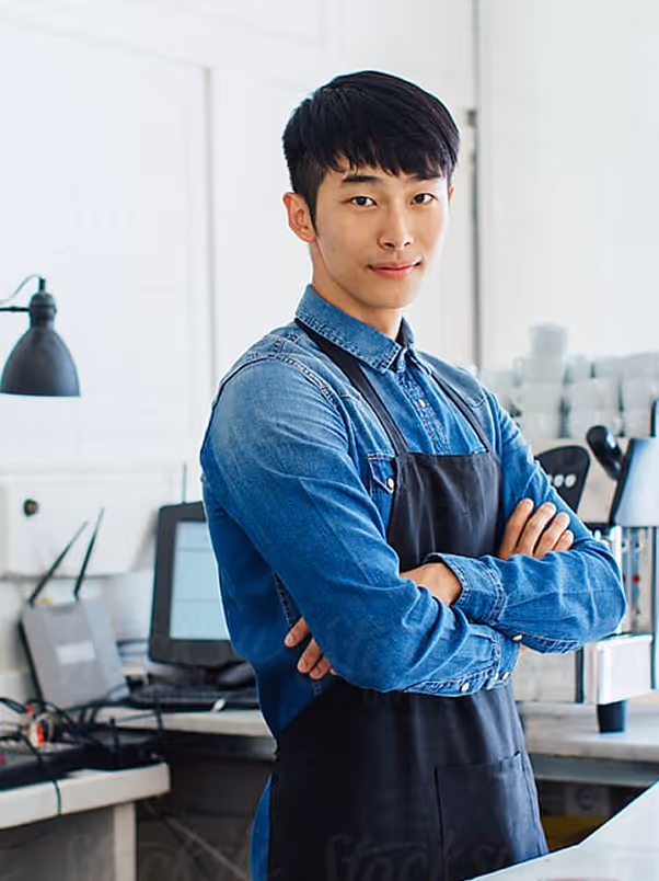 Barista wearing denim shirt and black apron standing with arms crossed in a coffee shop.