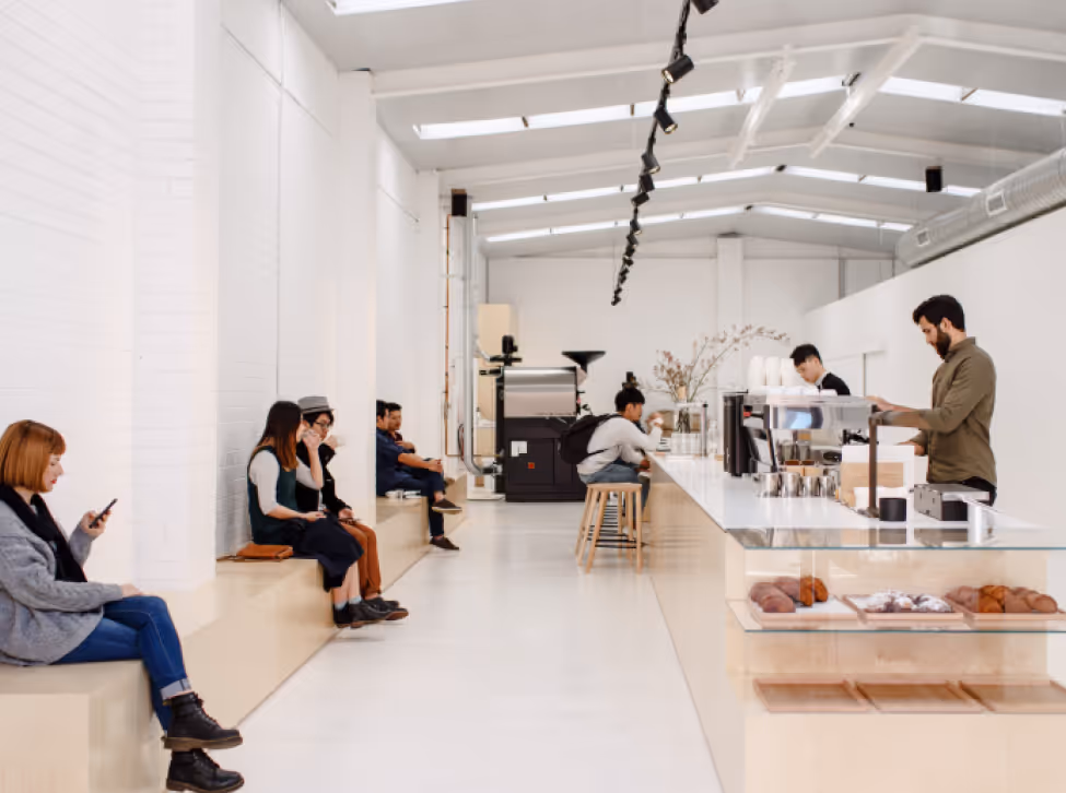 Modern minimalistic coffee shop interior with customers sitting on benches along the wall and baristas working behind a white counter displaying pastries.