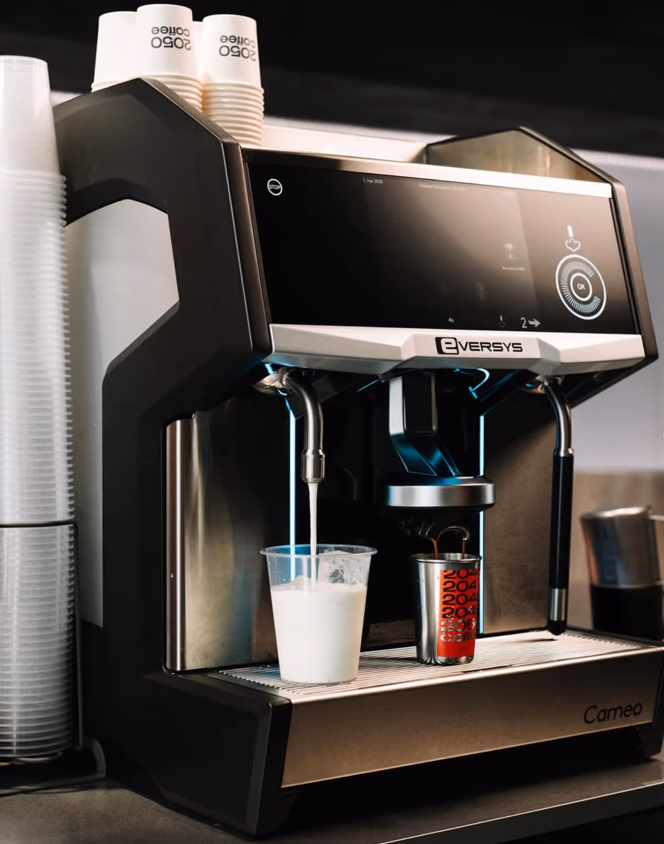 Eversys Cameo coffee machine dispensing milk into a plastic cup and espresso into a small metal cup on a countertop.