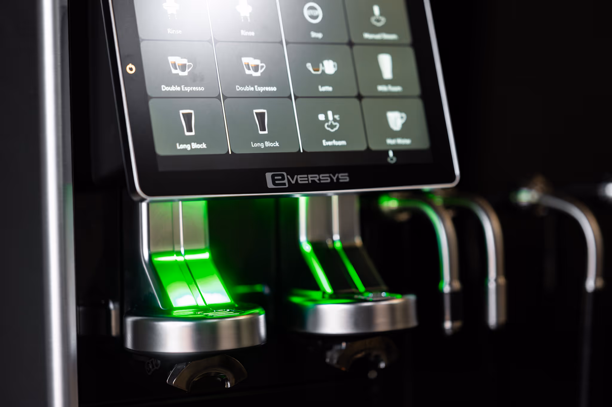 Close-up of an Eversys coffee machine touchscreen displaying coffee options with illuminated green dispensing nozzles below.