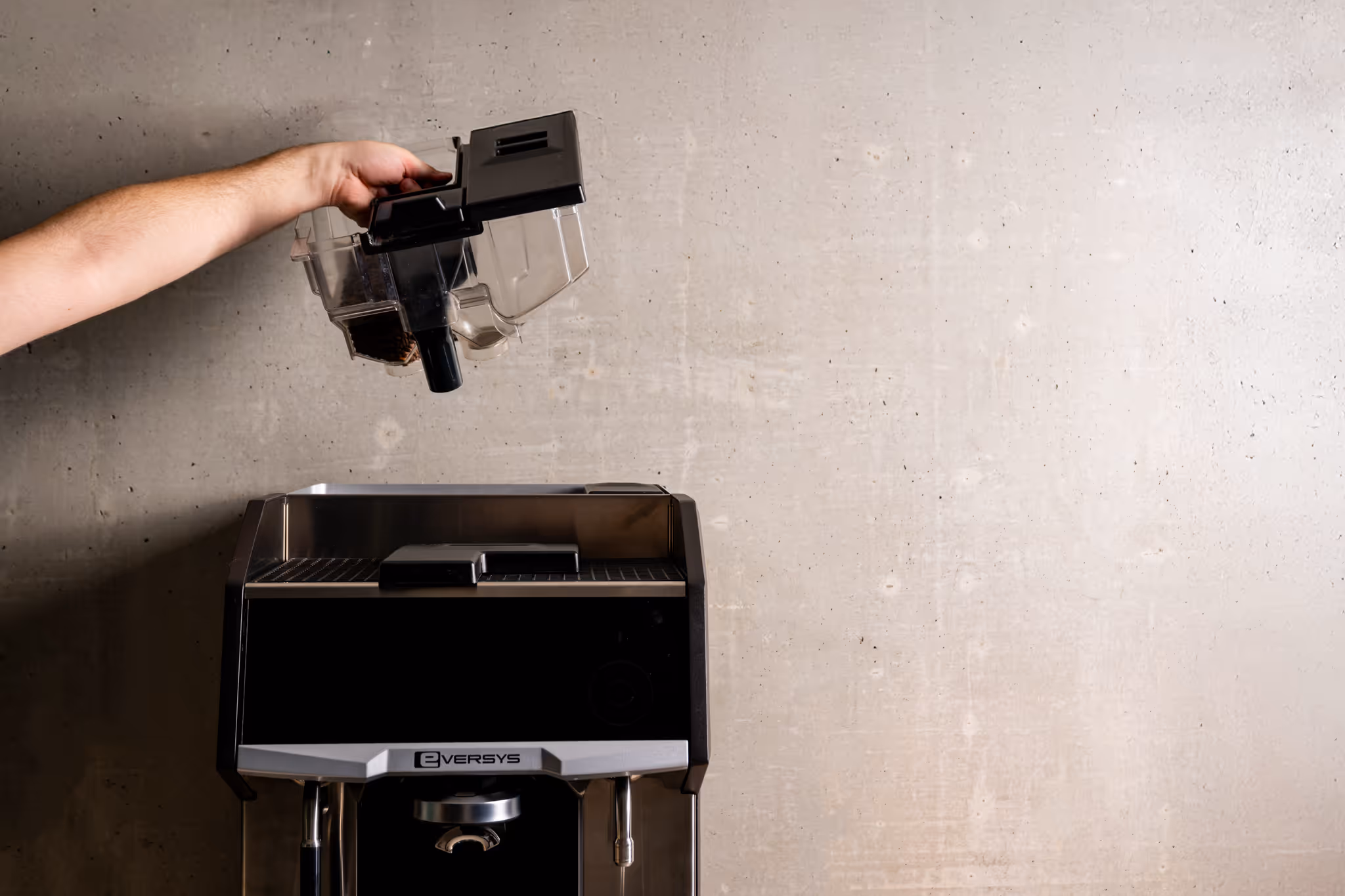 Hand holding a transparent coffee bean container above an Eversys coffee machine against a concrete wall background.
