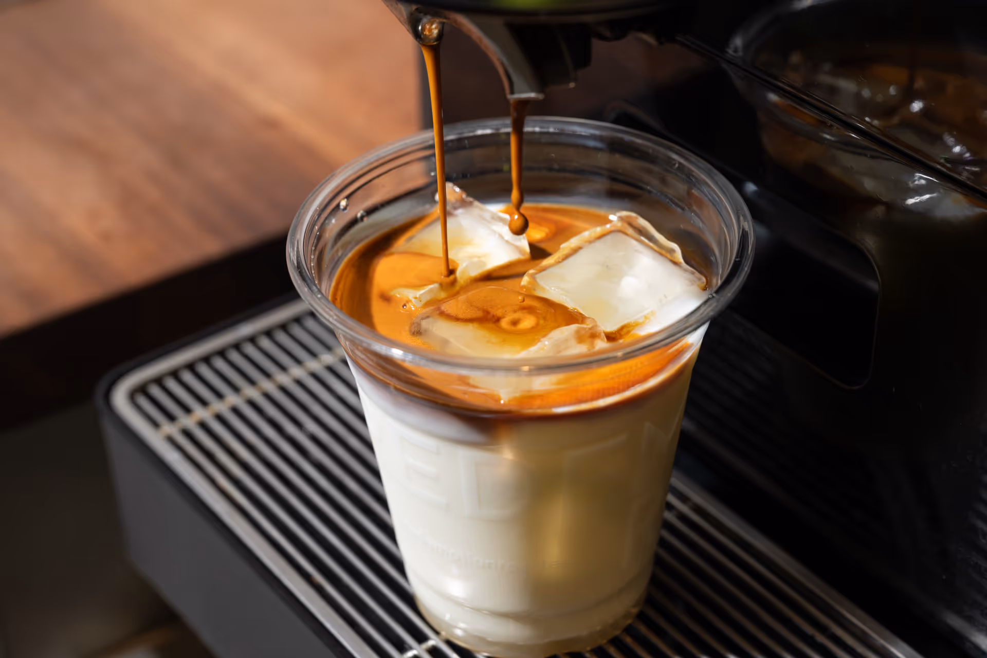 Espresso pouring over ice cubes in a clear plastic cup filled with milk on a coffee machine drip tray.