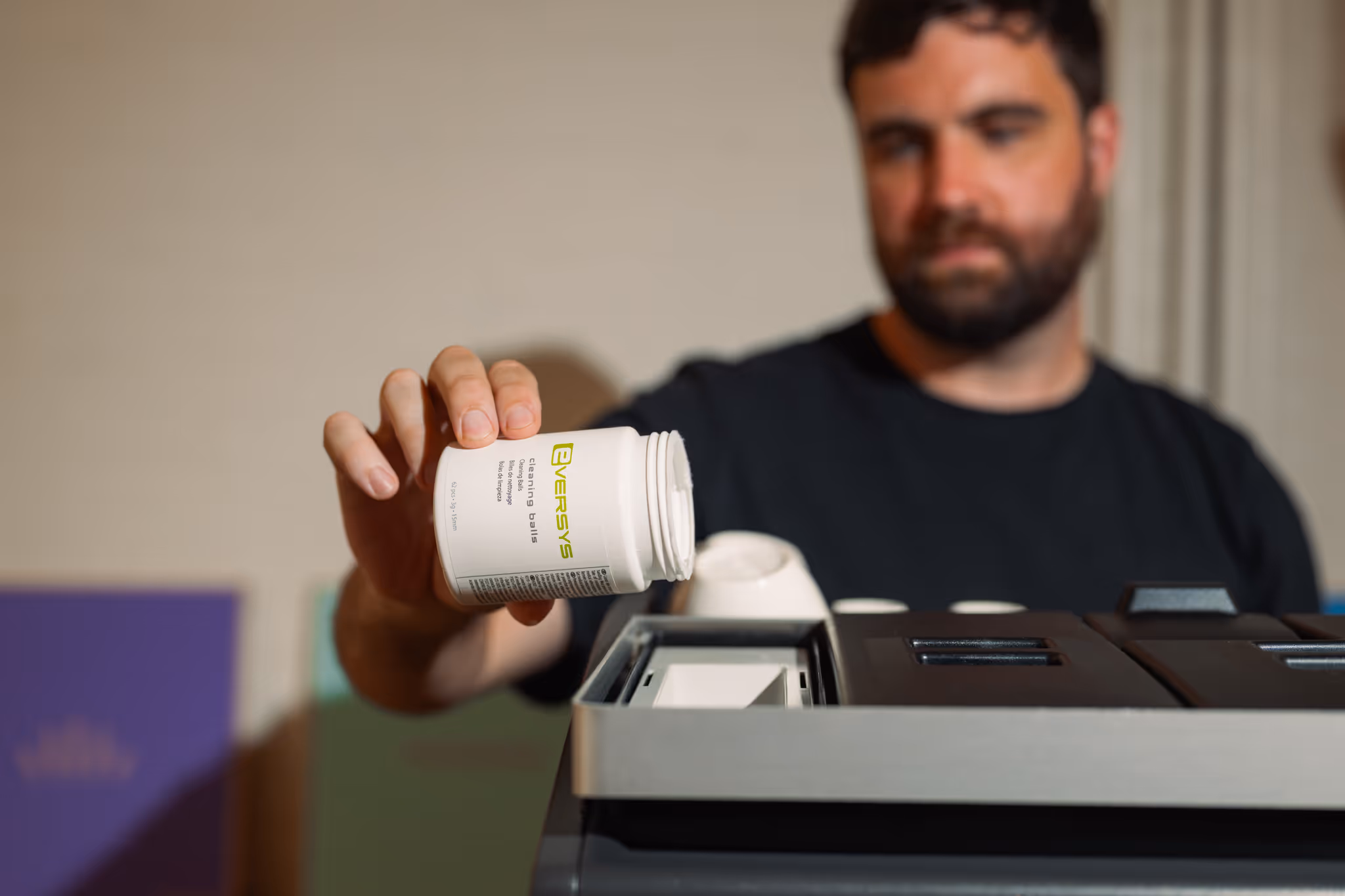Person wearing a white t-shirt with a green cartoon computer and 'Out of Office' text standing next to a modern Eversys Cameo coffee machine with cups on top.