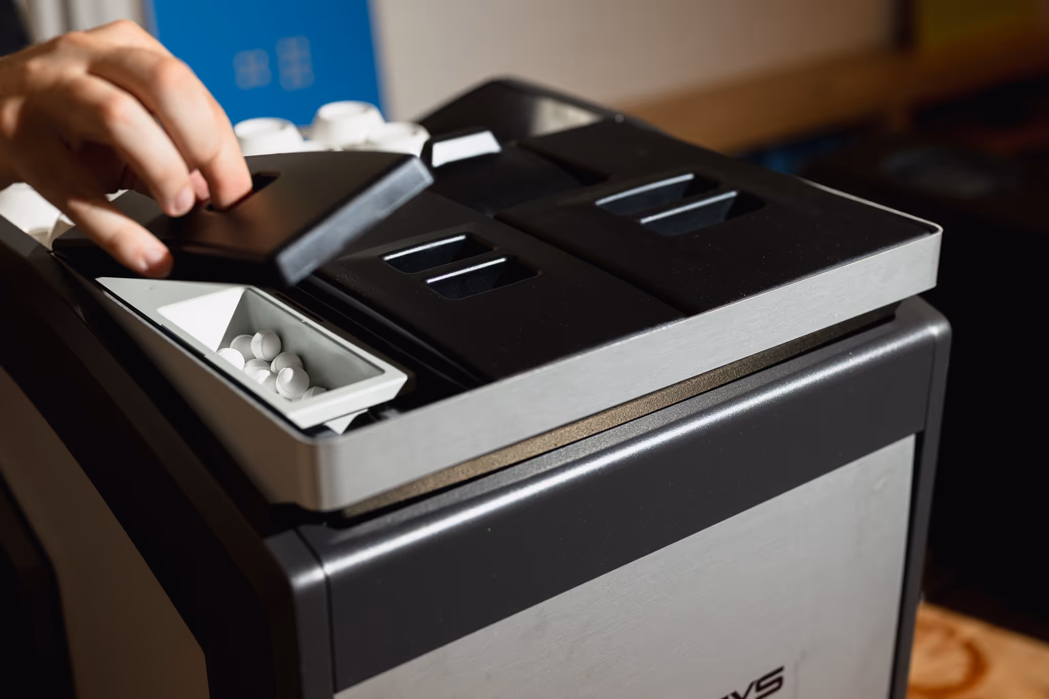 Hand removing the lid of a compartment filled with white round coffee capsules on a modern coffee machine.