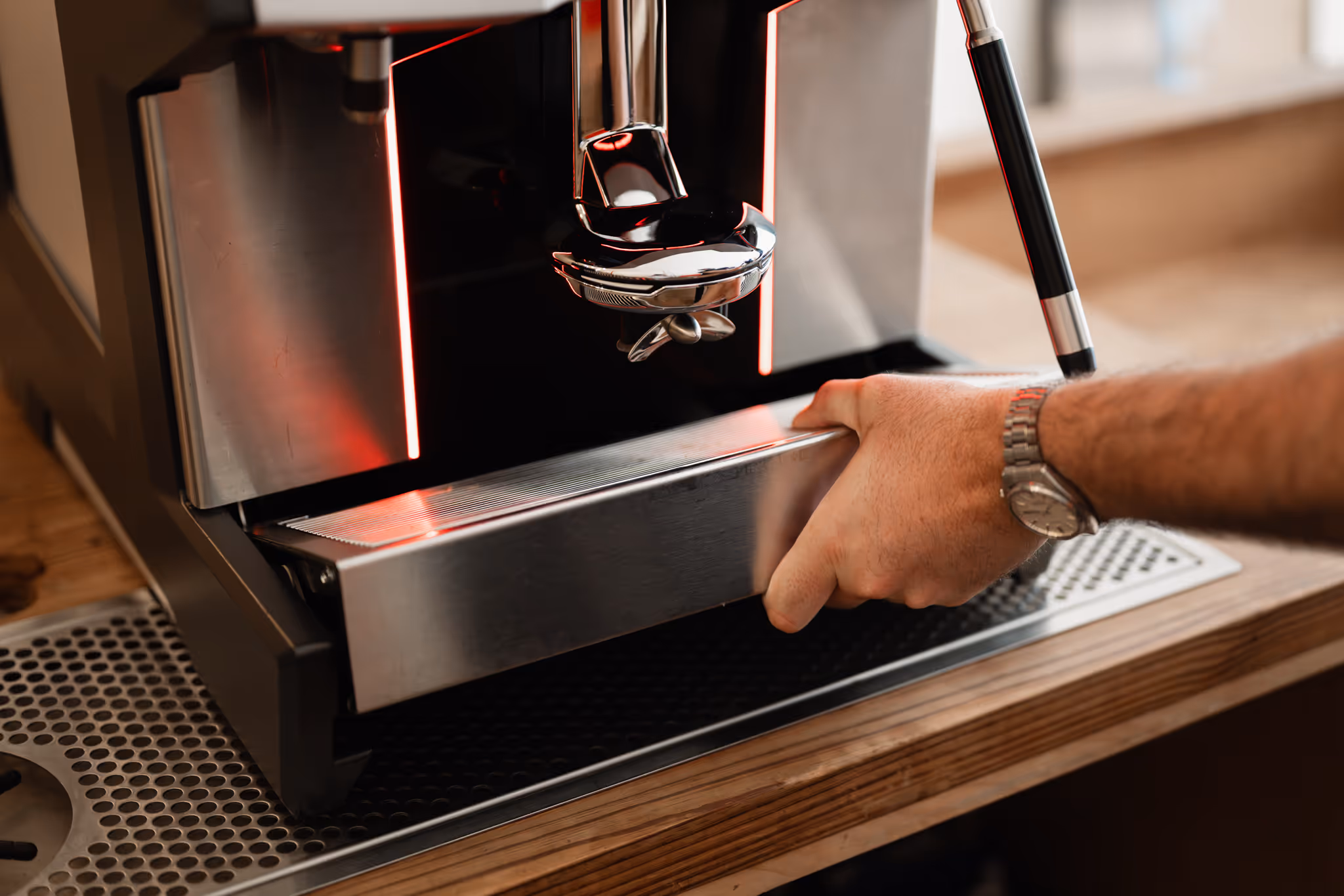 Person's hand pulling out the drip tray of a modern espresso machine on a wooden counter.