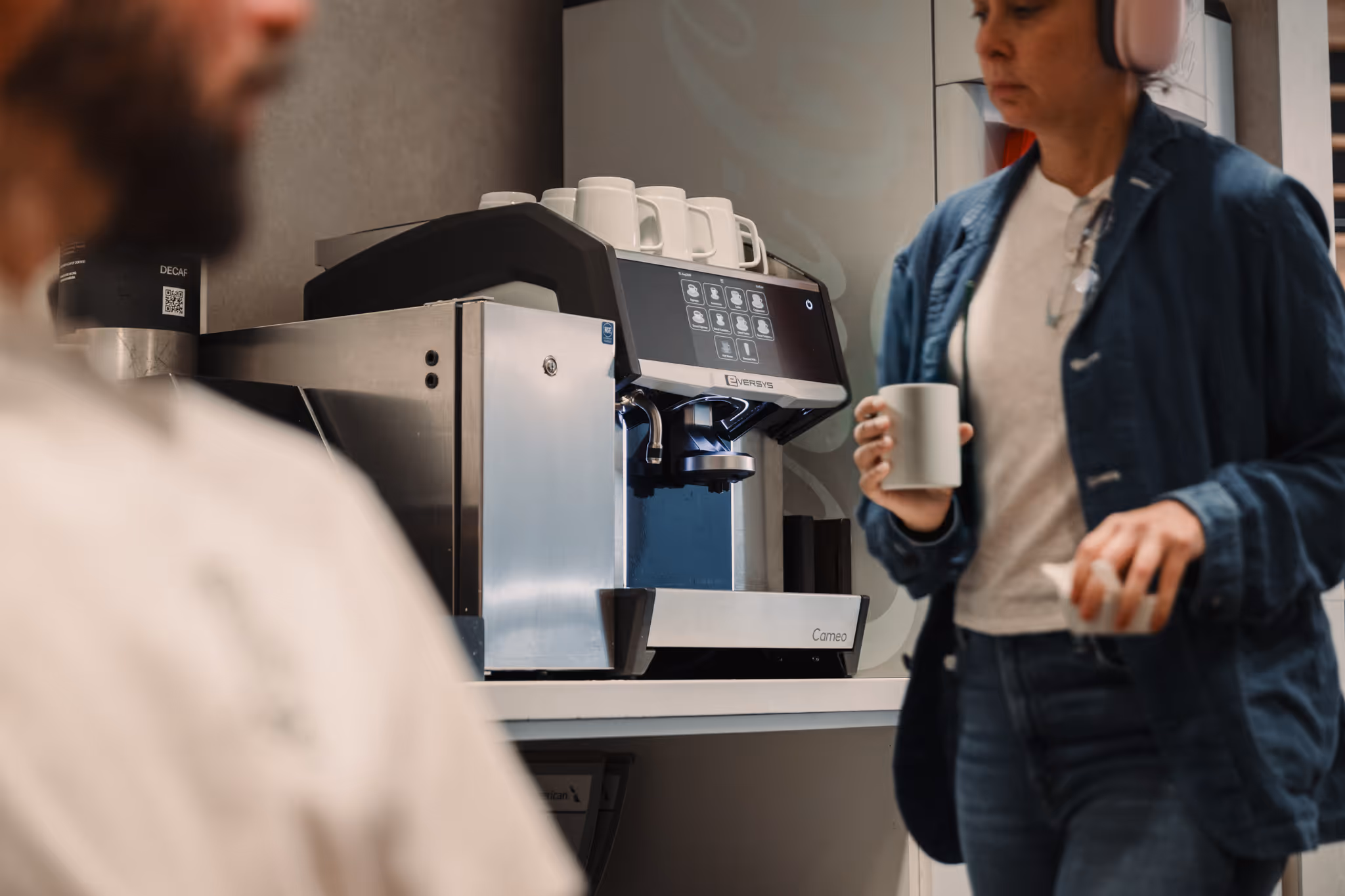 Modern coffee machine with stacked cups on top, a person holding a mug in the foreground.