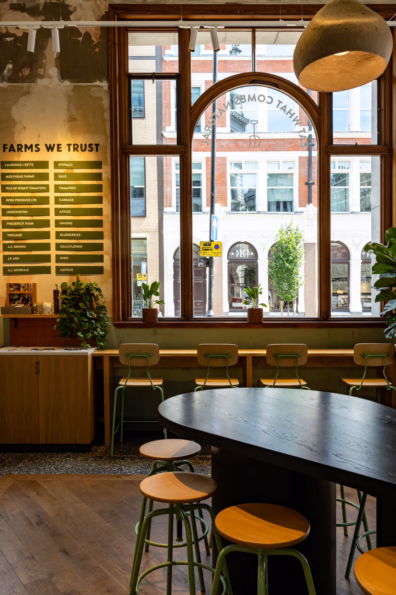 Cozy cafe interior with wooden stools around a dark round table, green chairs by a window, plants, and a wall listing trusted farms and their produce.