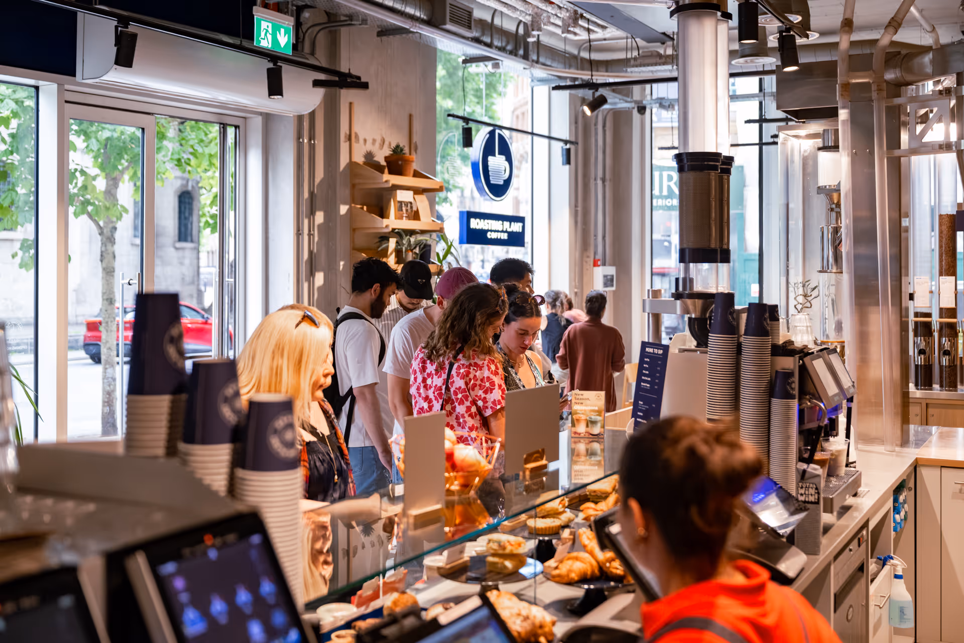 Customers waiting in line and a barista working behind the counter in a busy modern coffee shop.
