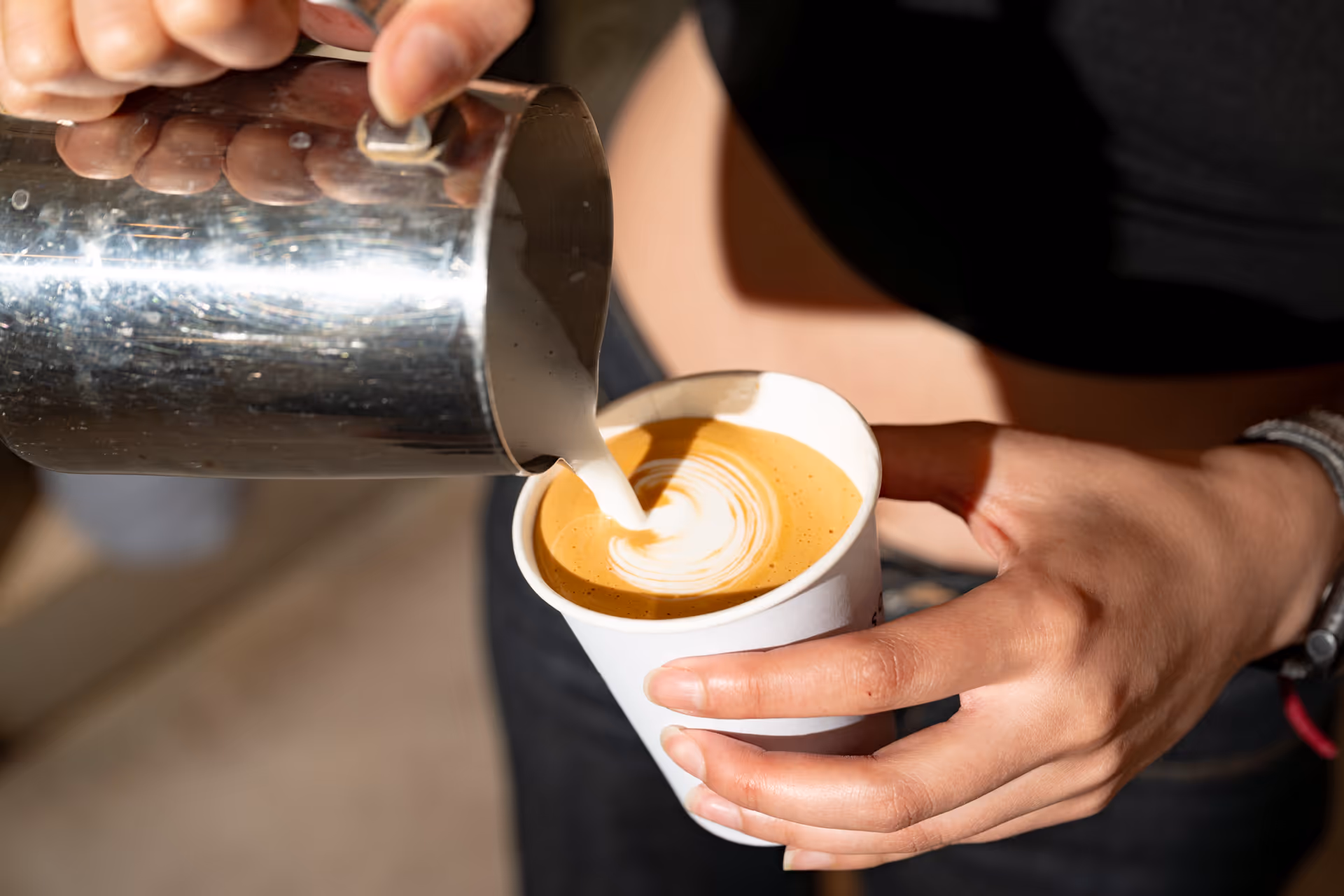 Hand pouring steamed milk from a metal pitcher into a paper cup of coffee to create latte art.