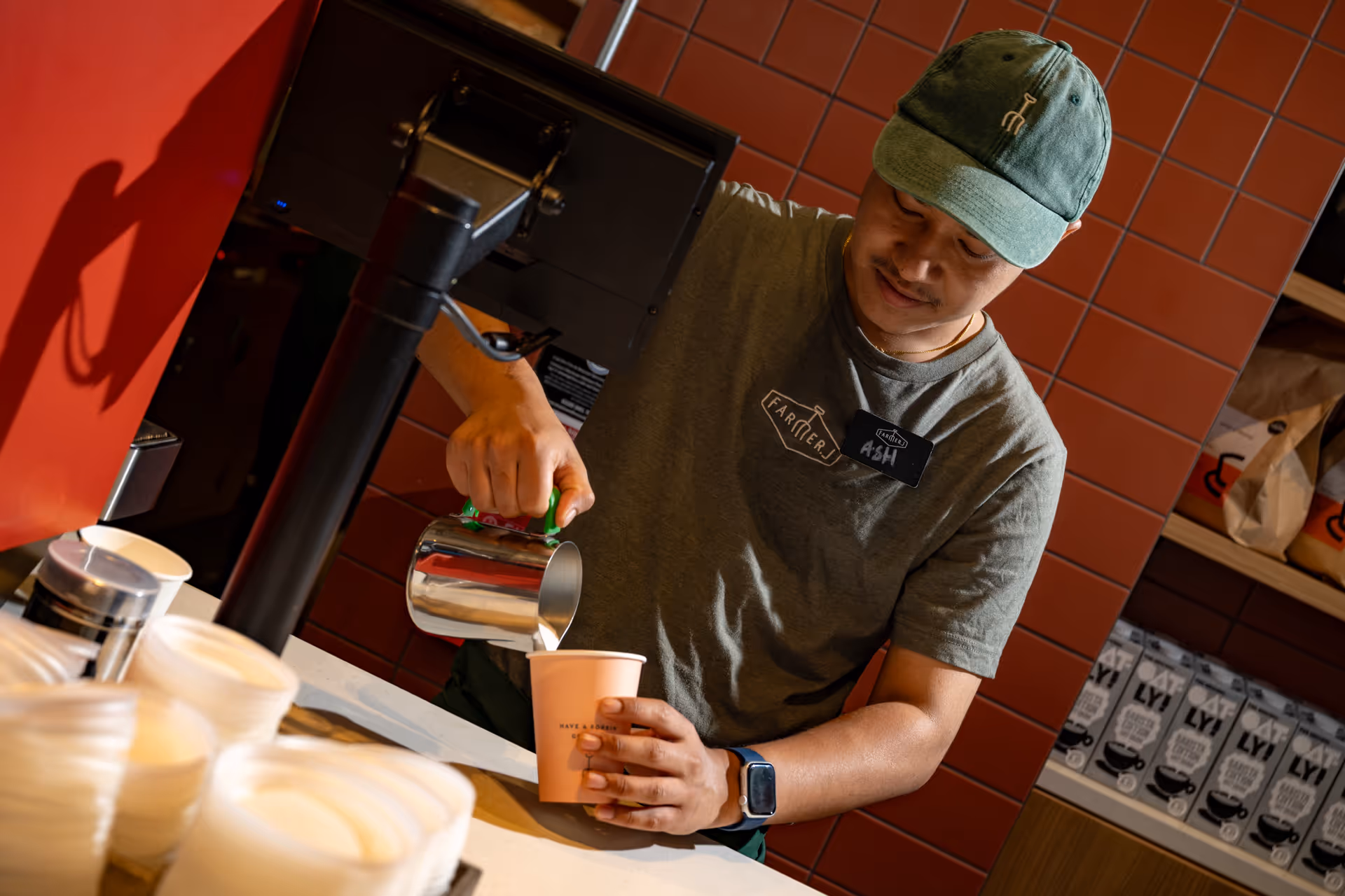 Barista wearing green hat and t-shirt pouring milk into a pink cup at a coffee counter.