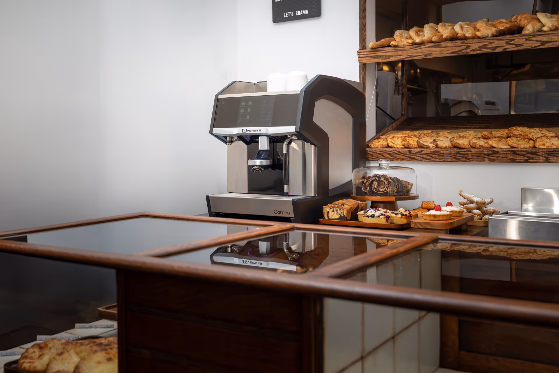 Modern Eversys Cameo coffee machine on a counter next to shelves with assorted breads and pastries in a bakery.
