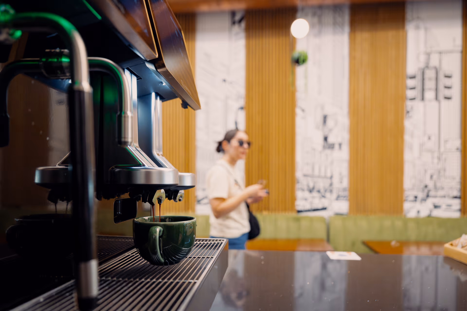 Coffee machine dispensing espresso into a green cup with a person blurred in the background inside a café.