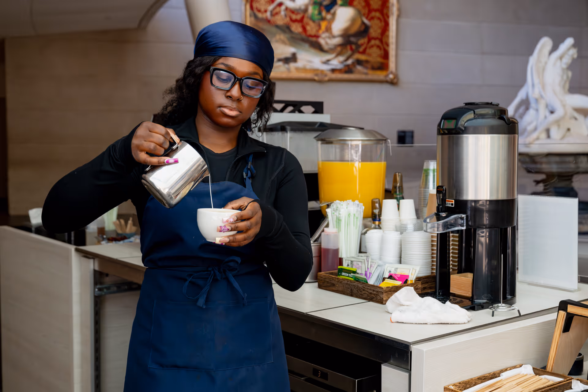 Barista in navy apron pouring milk into a white cup behind a beverage station with juice dispenser and coffee urn.