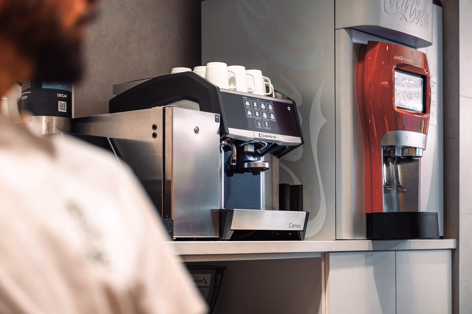 Commercial coffee machine with cups on top beside a Coca-Cola Freestyle dispenser on a countertop.
