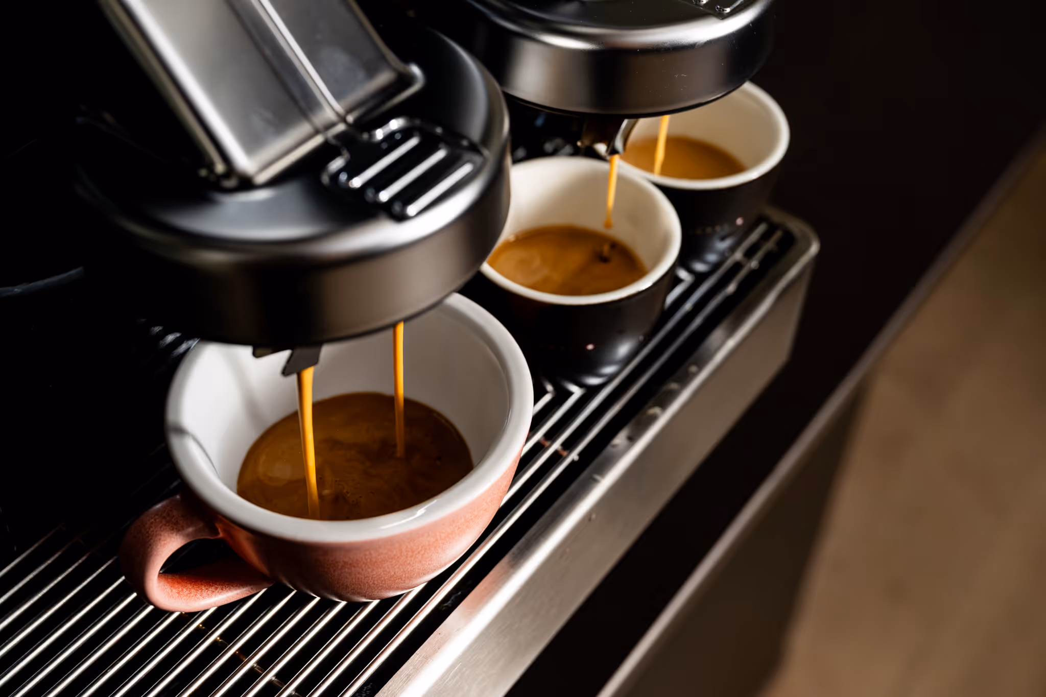 Close-up of espresso pouring from a coffee machine into two black cups and one pink ceramic cup.