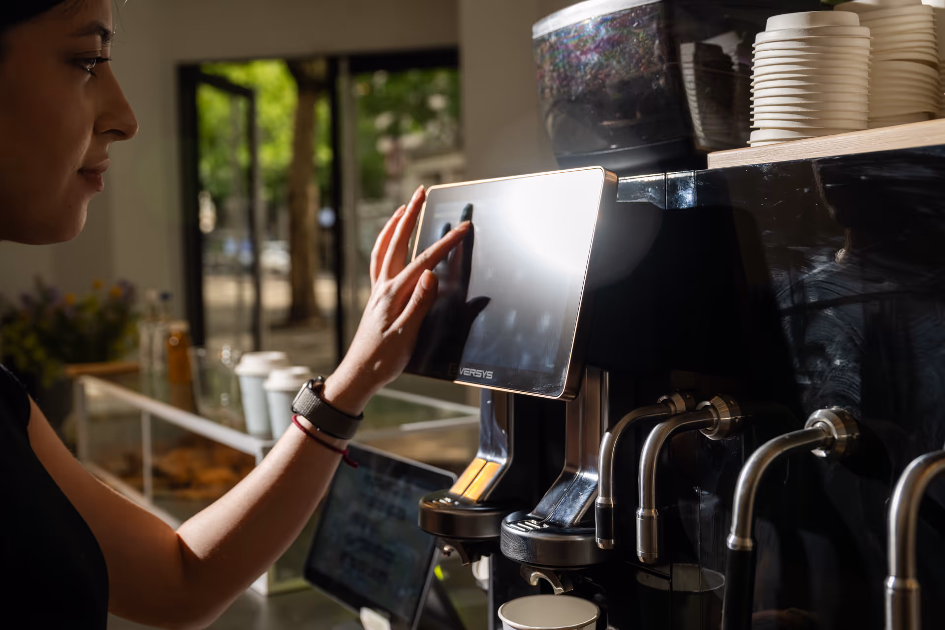 Woman operating a touchscreen on a commercial coffee machine with cups stacked on top.