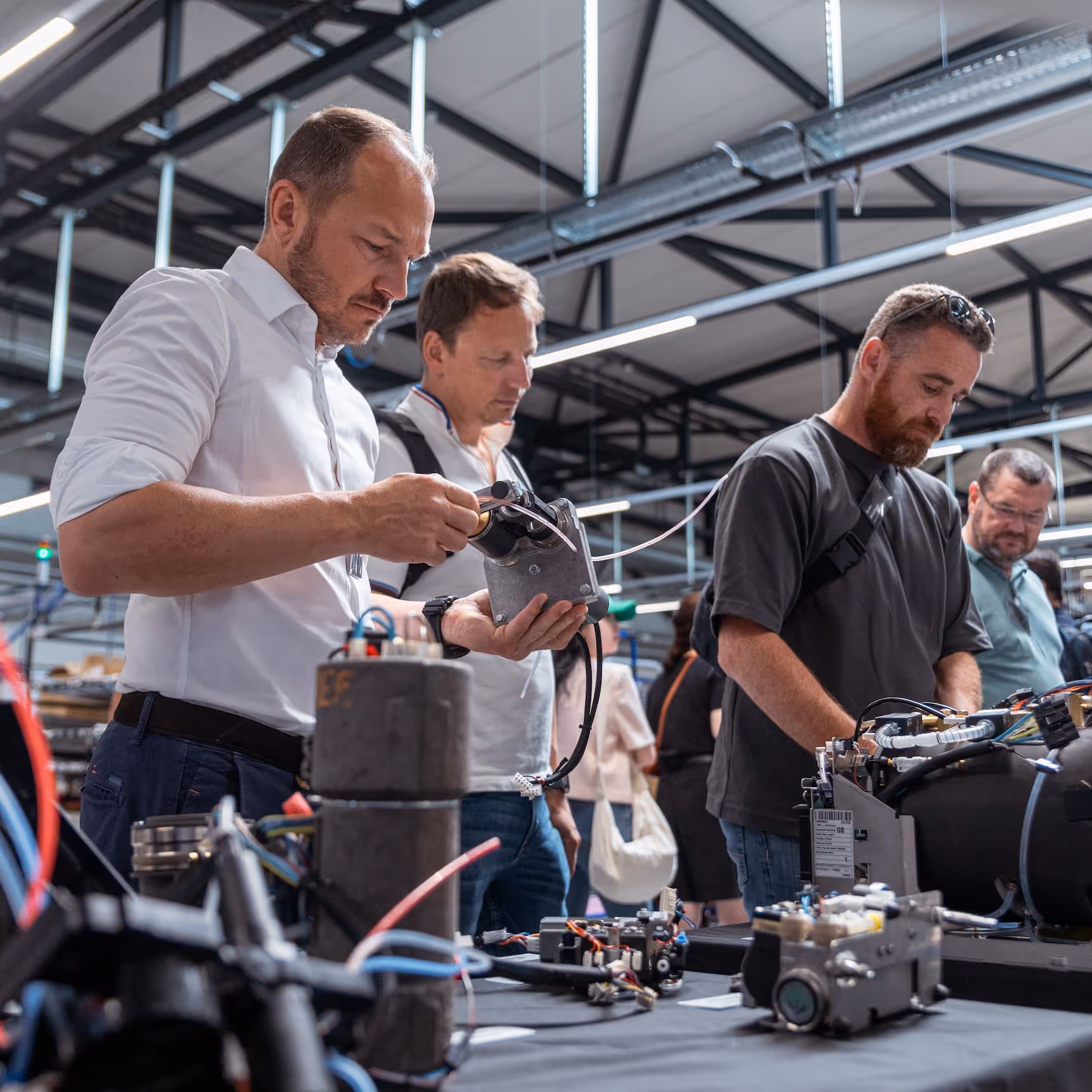 Three men examining electronic components and machinery parts on a table inside an industrial workshop.