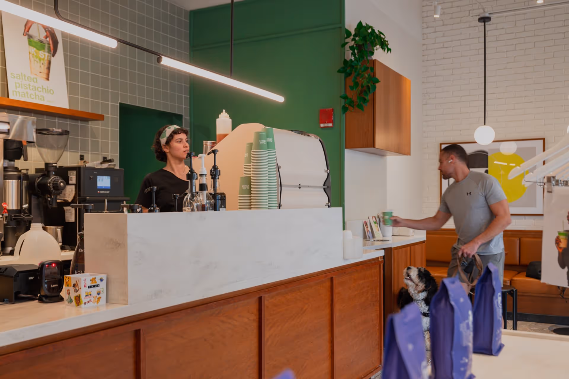 Barista stands behind a coffee counter with stacked green cups, while a man in gray athletic wear holds a cup and a dog looks up at him in a modern cafe.