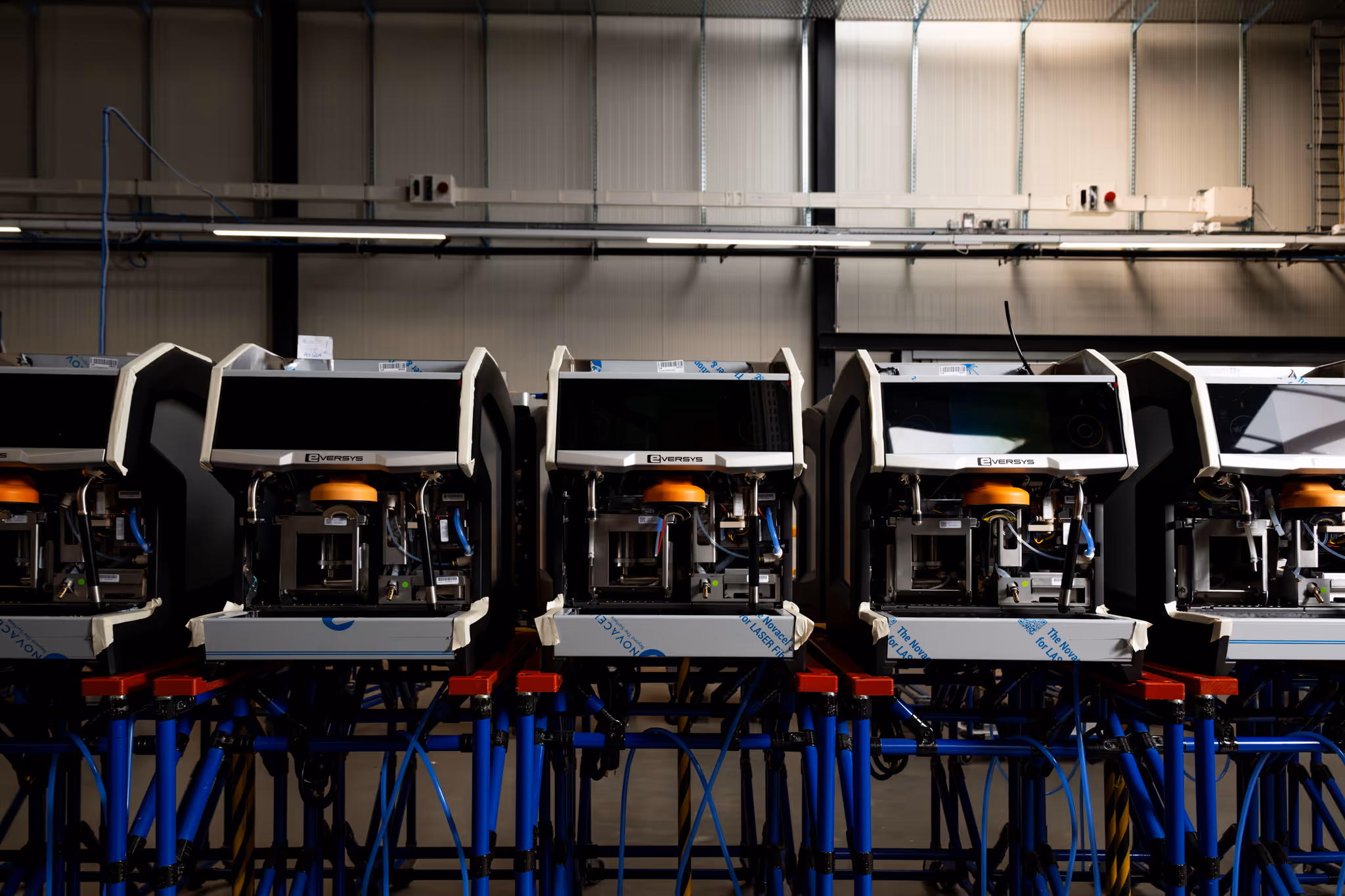 Row of Eversys branded coffee machines on blue and red metal stands inside a production facility.