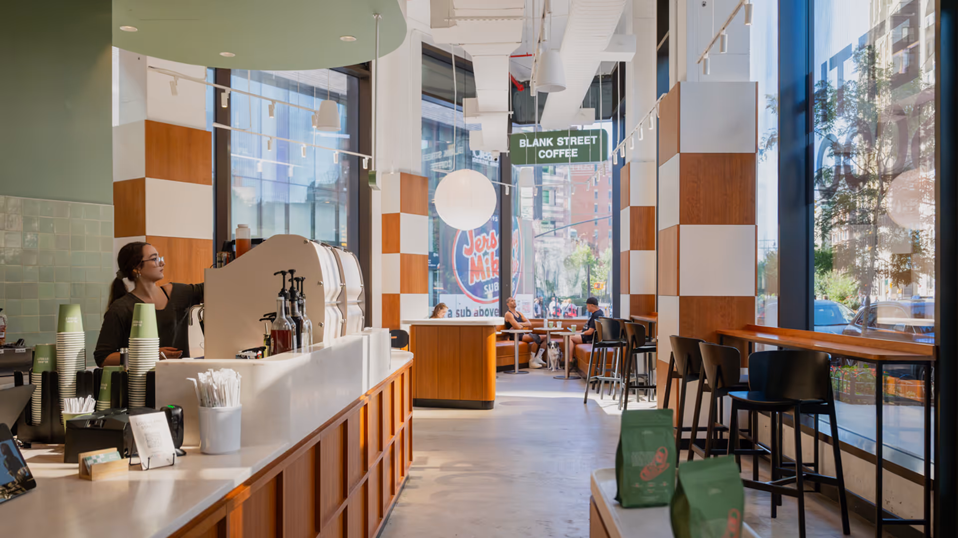 Modern coffee shop interior with barista behind counter, wooden decor, and customers sitting at tables with a dog.