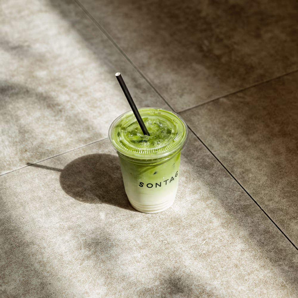 Iced matcha latte in a clear plastic cup with a black straw casting a shadow on tiled floor.