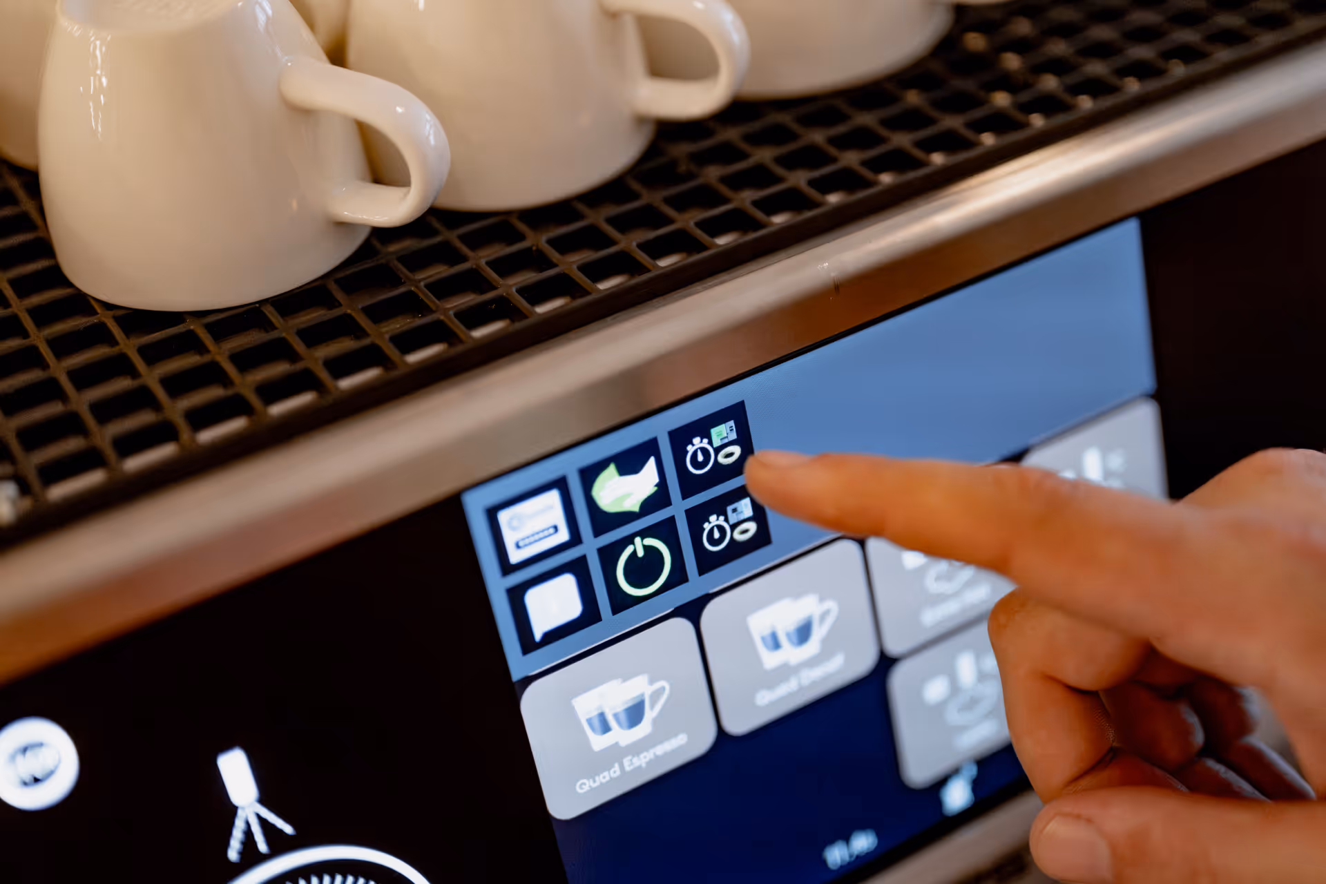Close-up of a hand touching the touchscreen interface of a coffee machine with white cups on top.