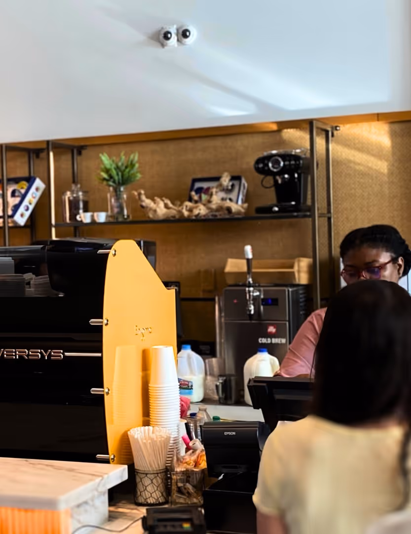 Barista working behind a coffee counter with espresso machine, cups, and coffee supplies, serving a customer.