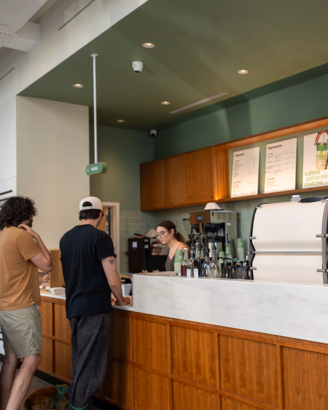 Barista with a tattoo operating a touchscreen register in a modern café with coffee machines, pastries, and customers in the background.