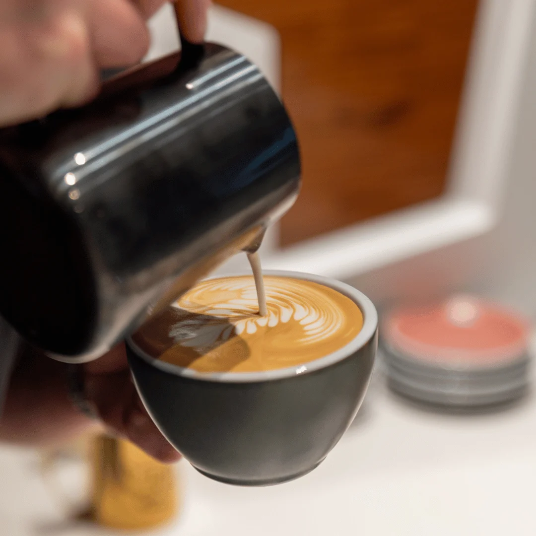 Person pouring steamed milk from a metal pitcher into a cup of coffee creating latte art.