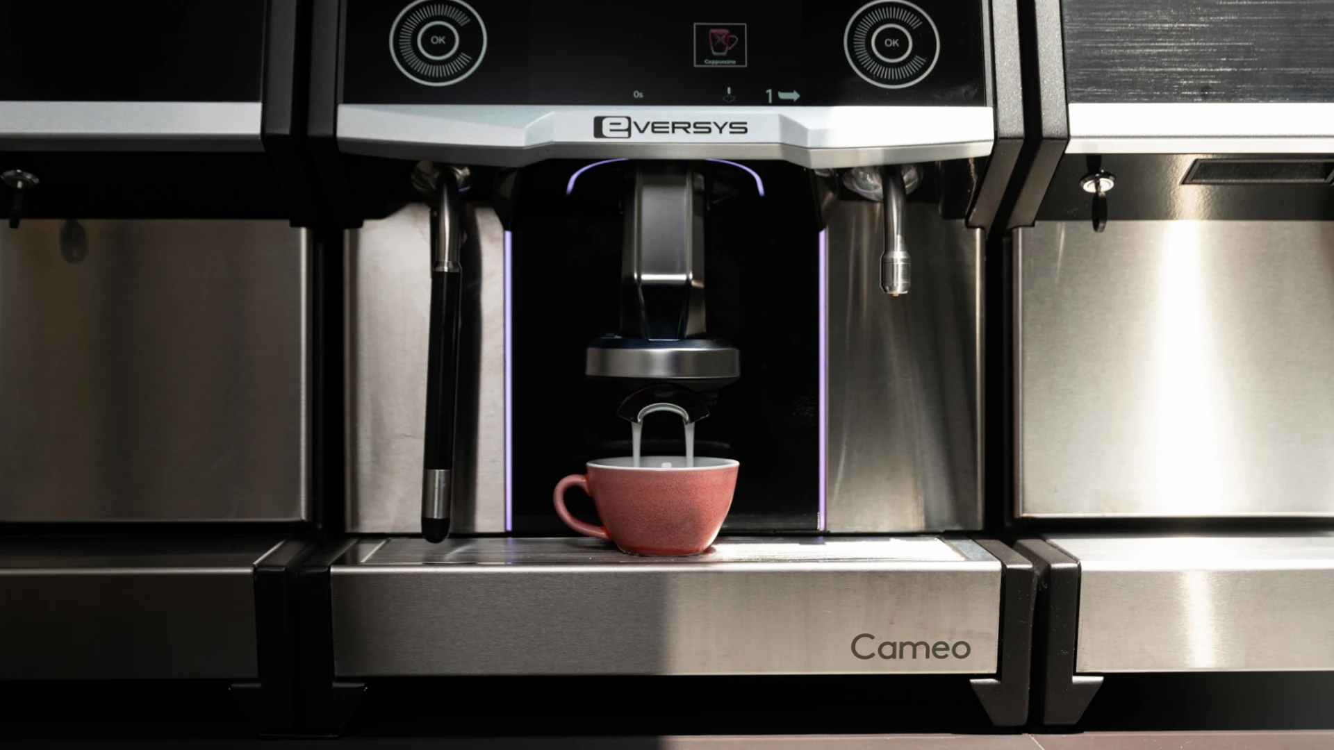 Barista in black apron pouring steamed milk from a metal pitcher into a gray ceramic cup next to a coffee machine.