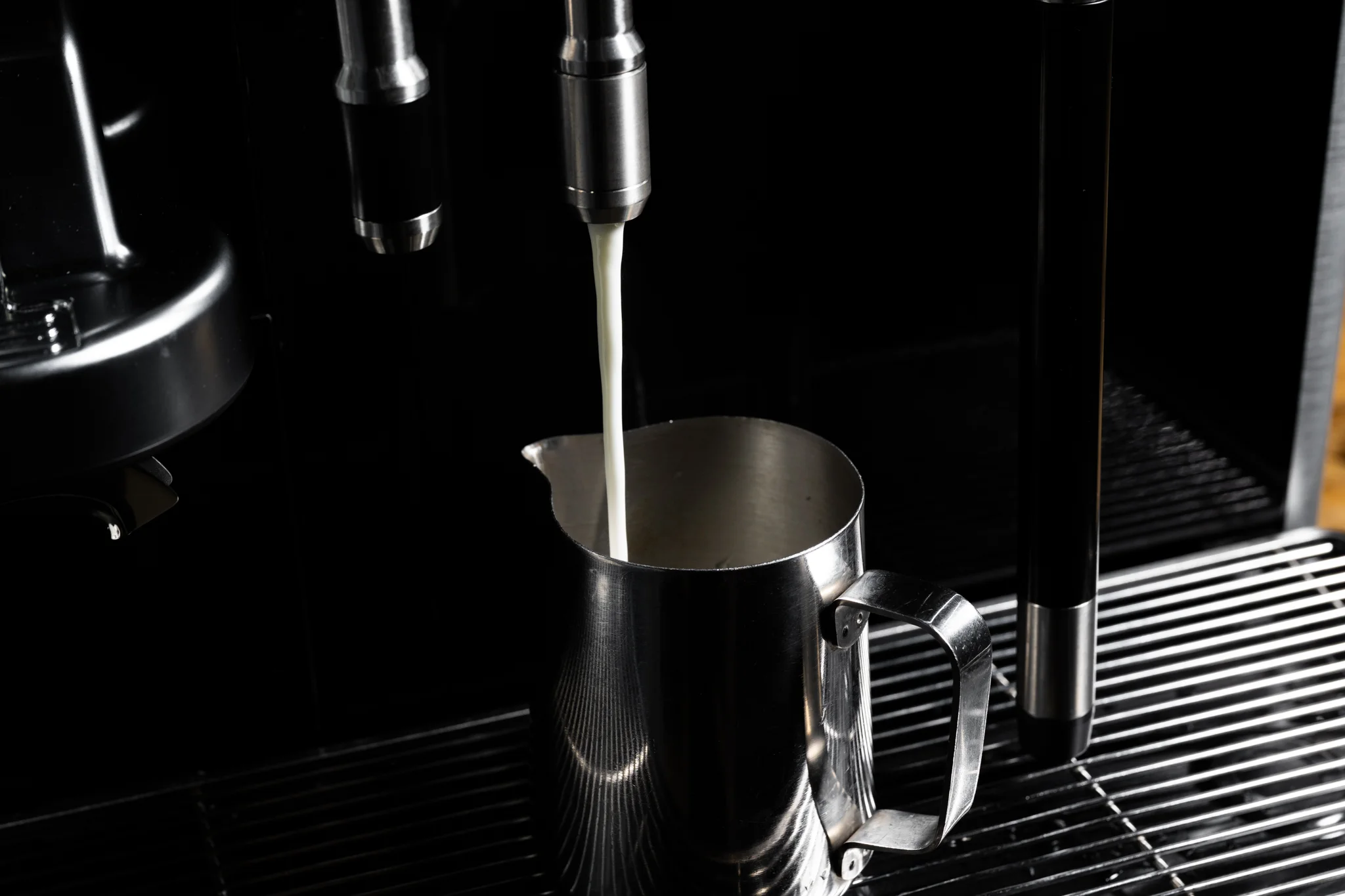 Coffee machine pouring steamed milk into a stainless steel frothing pitcher.
