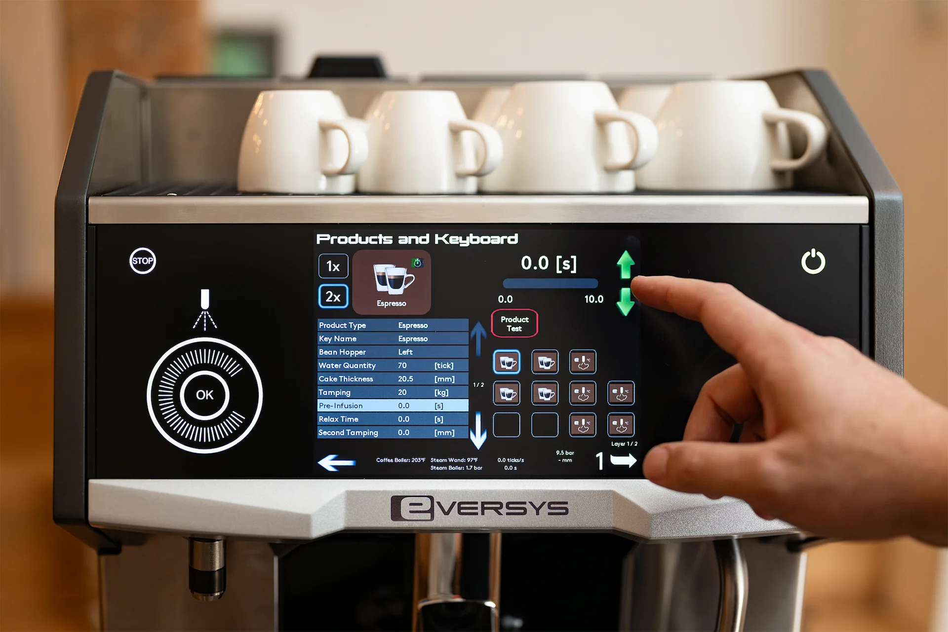 Close-up of a hand adjusting settings on an Eversys espresso machine touchscreen display with white cups on top.