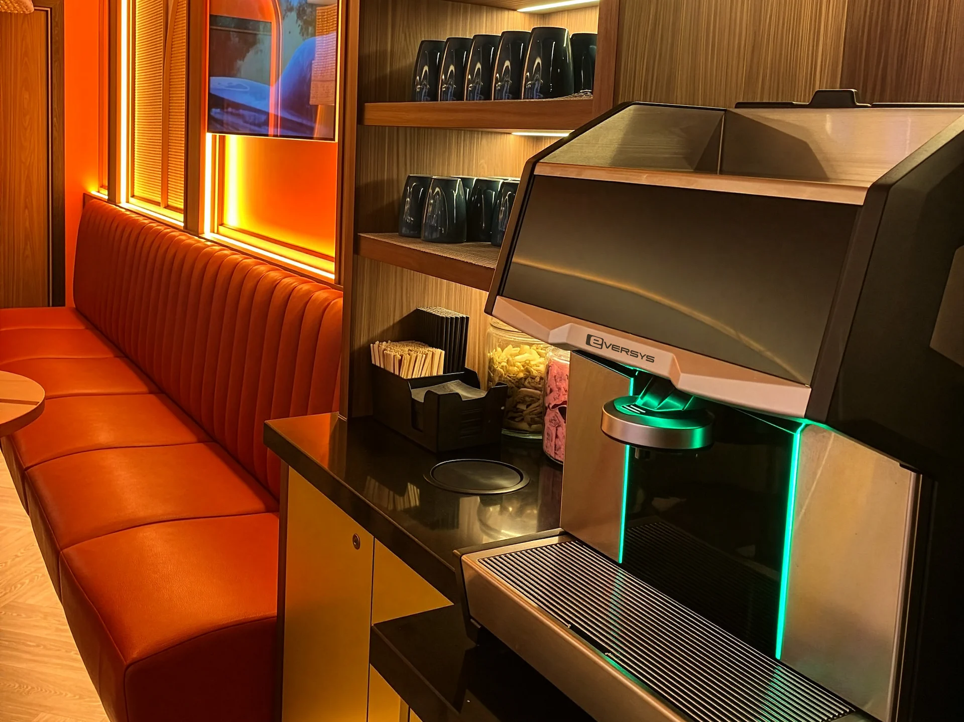 Person wearing a white t-shirt with a green 'Out of Office' graphic standing next to an Eversys Cameo coffee machine with cups on top.