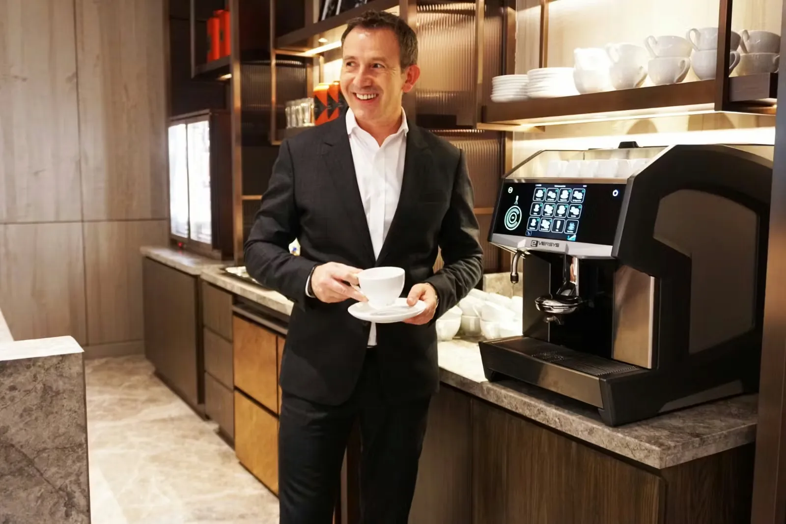 Woman holding a white cup and coffee pods near an Eversys Cameo coffee machine with stacked cups on top.