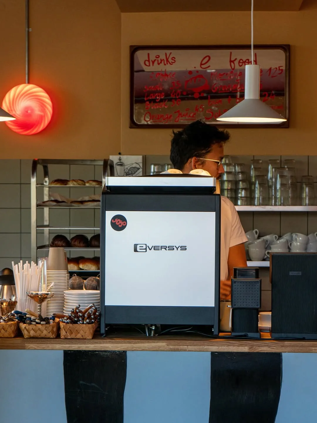 Close-up of an Eversys espresso machine dispensing coffee into a blue Roasting Plant Coffee cup, with stacks of coffee cups in the background.