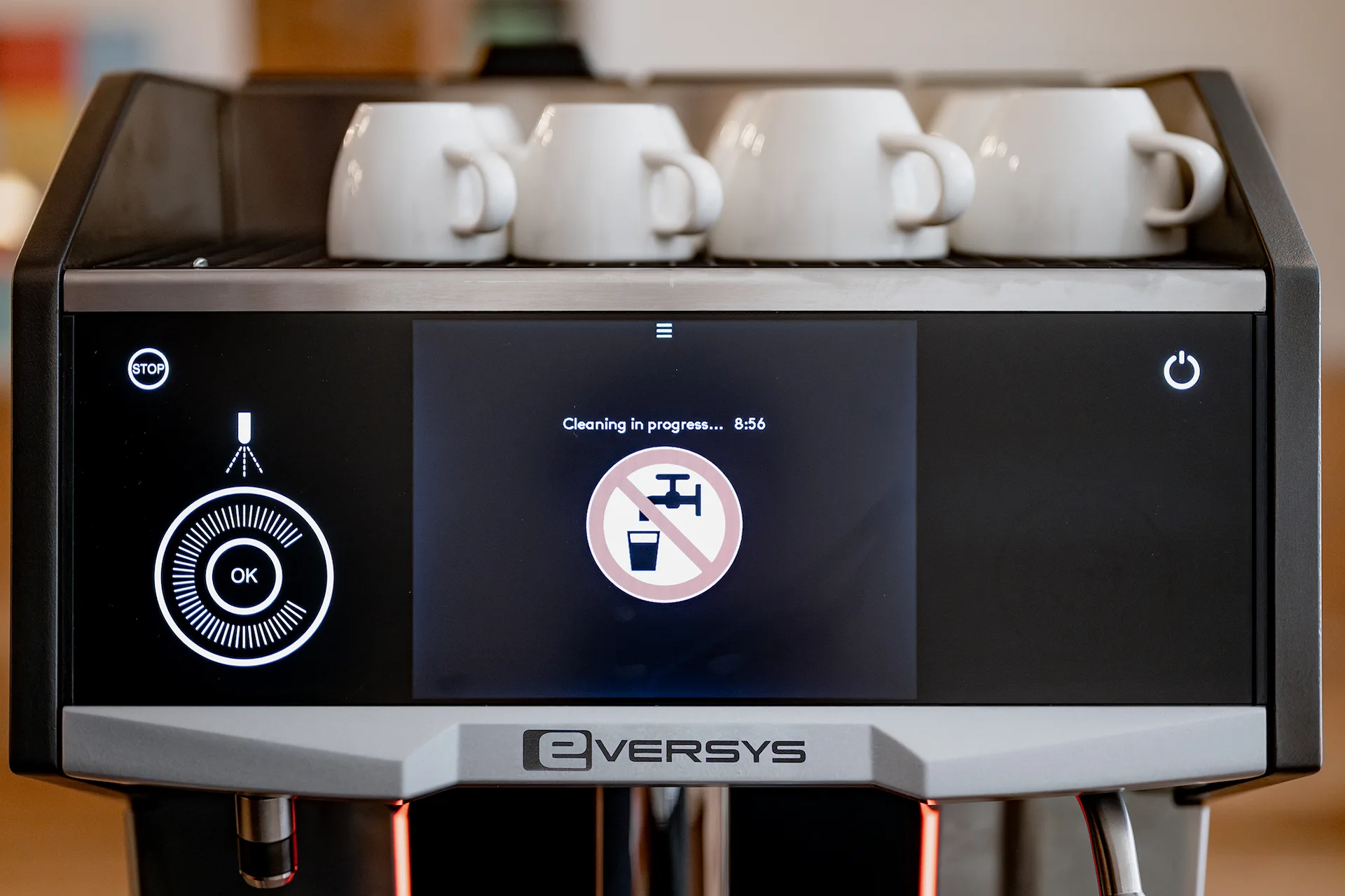 Eversys coffee machine display showing cleaning in progress with a no drinking water icon and four white coffee cups on top.