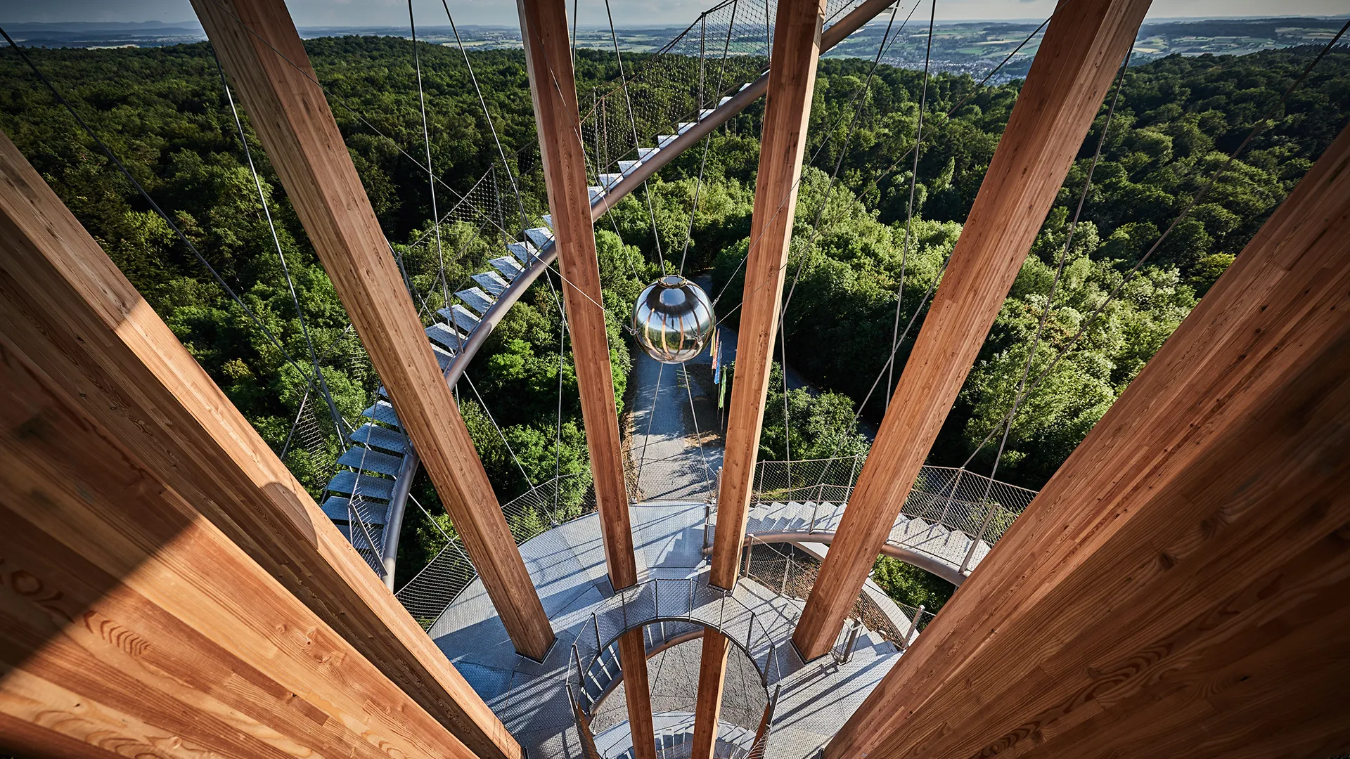 Blick von einem hölzernen Aussichtsturm auf eine spiralförmige Treppe und dichten grünen Wald darunter.