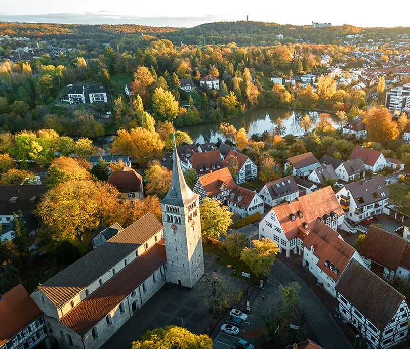 Luftaufnahme einer historischen Kirche mit spitzem Turm umgeben von herbstlich gefärbten Bäumen und einem See in einer Stadt.